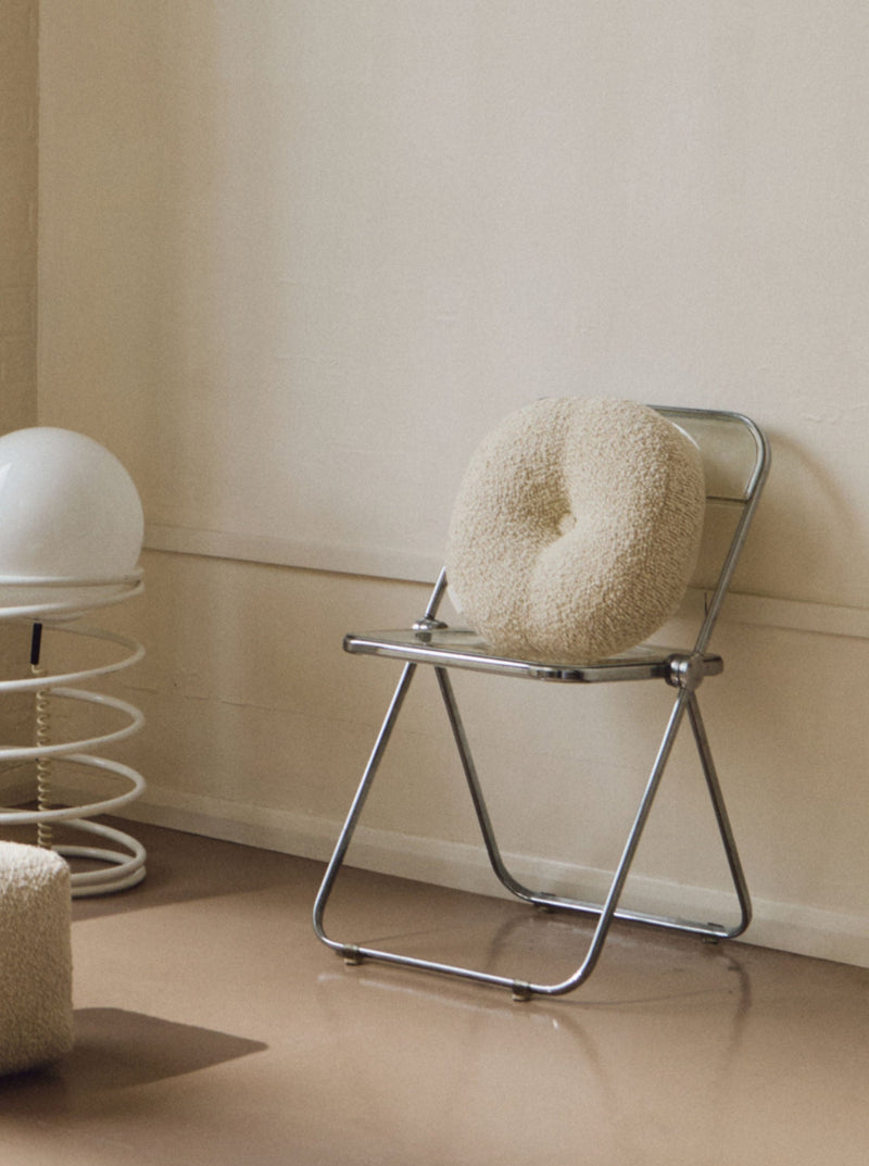 A minimalist room featuring a metal folding chair with a handmade cushion made from British eco wool, a spring-like white lamp with a round top, and Le Rond - Moyen - Wool Bouclé - Egg White by AMÉLIORÉ. The space has a neutral color palette with beige walls and a reflective, polished floor.