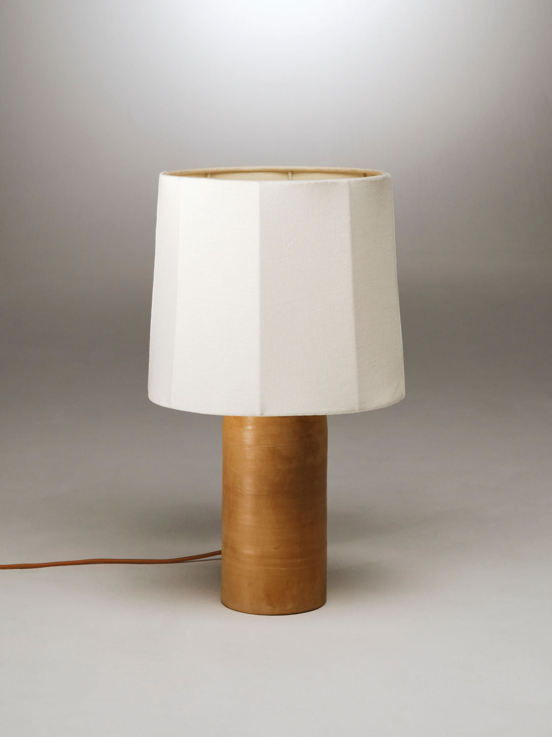 A Sonora Linen Table Lamp by Nassi Lamps with a cylindrical wooden base and a white, faceted fabric lampshade. Inspired by the charm of Puglia, the lamp's power cord extends from the base against a simple, neutral gradient background.