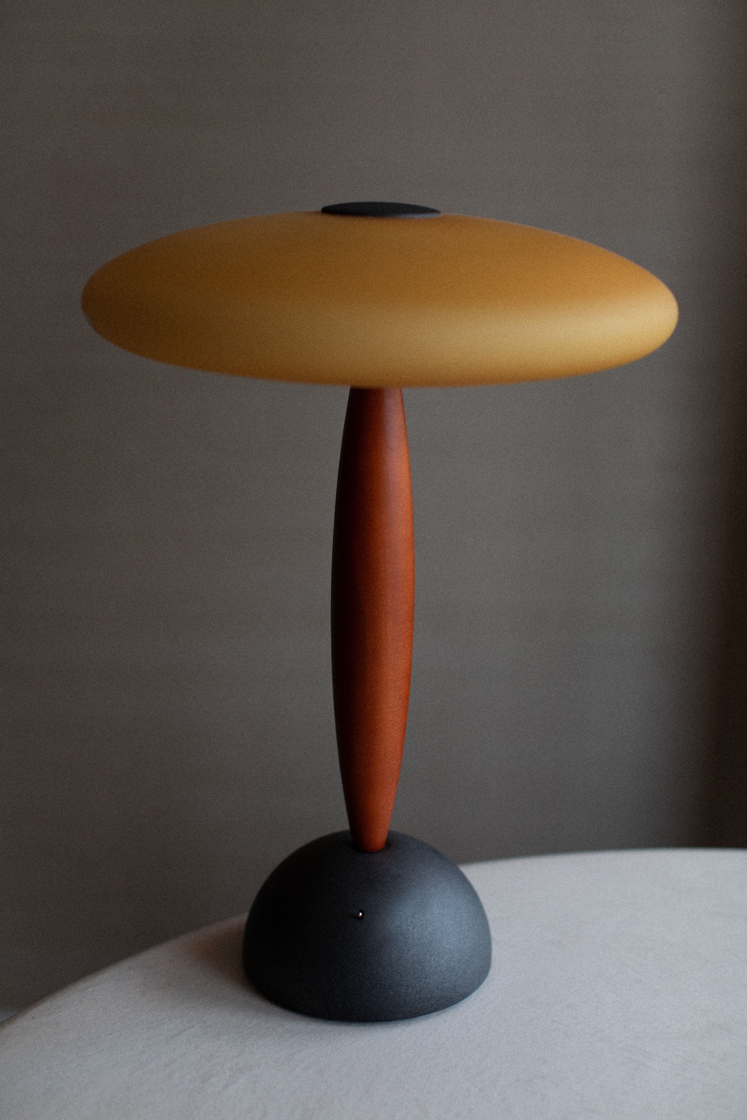 A contemporary handcrafted Vintage Murano Lamp by Out For Lunch with a unique design featuring a mustard yellow lampshade, a slender terracotta-colored stem, and a round matte black base, set against a neutral gray background.