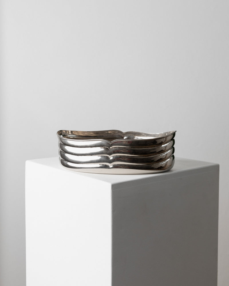 A Bowl Silver-plated - Italy 1960s by Bottega Jacobs rests on a white pedestal. Its wavy, multi-layered design elegantly reflects light, reminiscent of an elegant table decoration against a plain backdrop.