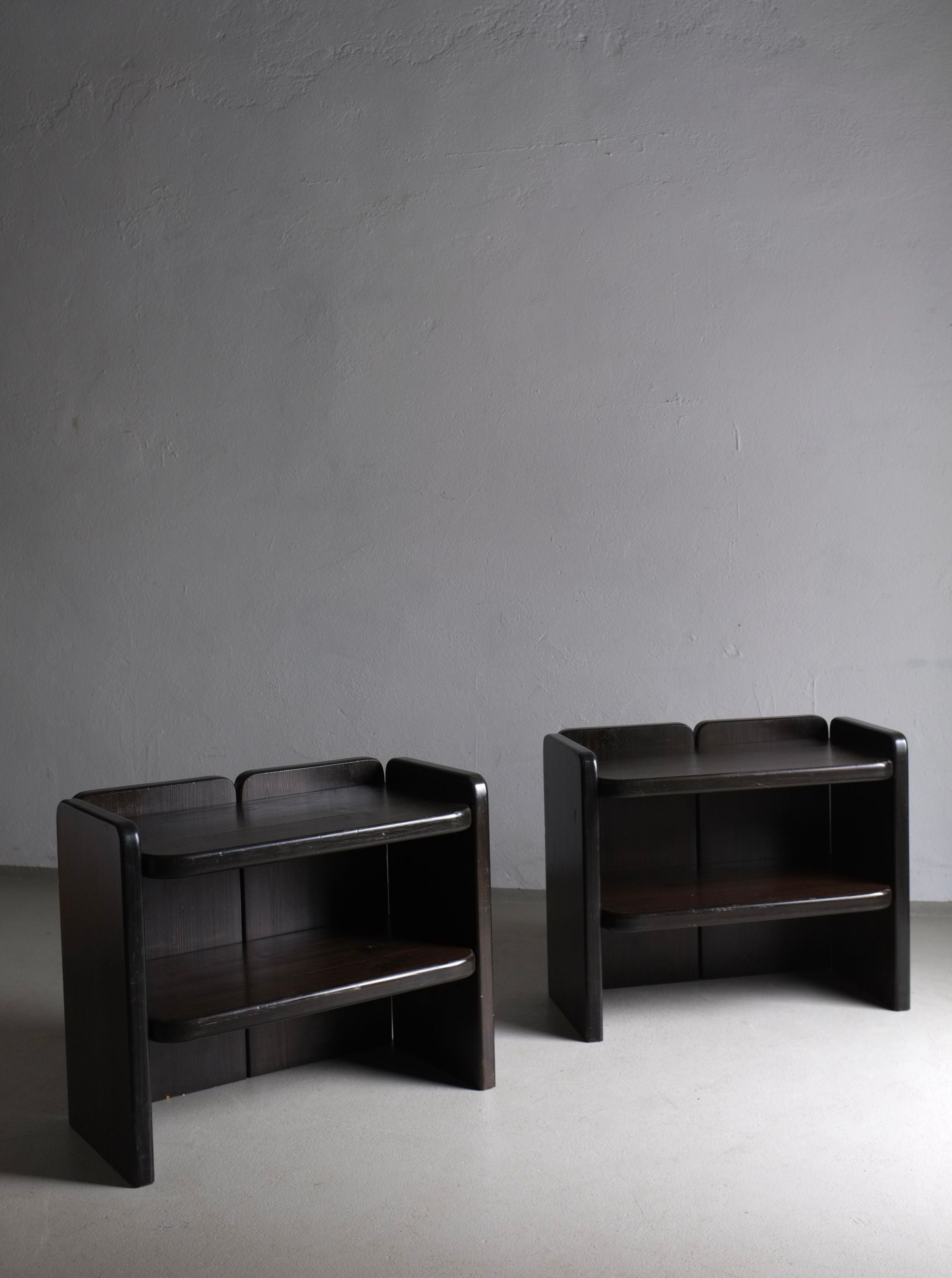 A minimalist composition featuring two identical *2 Brutalist Dark Wood Night Stands | 1970s* in vintage condition from *Veter Vintage*, with rounded corners and two shelves each, set against a plain, light grey wall and concrete floor. The setting is simple, highlighting the clean lines and functional design of the furniture.