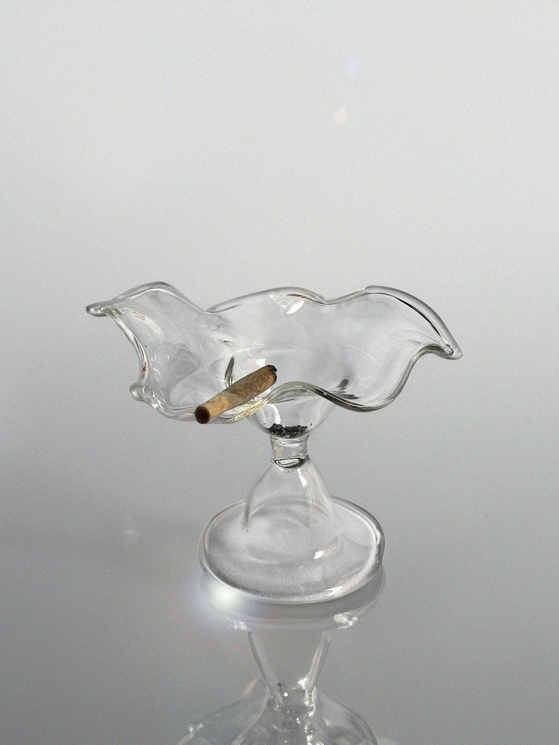 The 'Myth' Ashtray by Pia Glassworks is a handcrafted, pedestal-based glass piece with a wavy edge. It elegantly holds an unlit cigarette on its reflective surface and features a unique abstract design, perfect for decorative tableware.