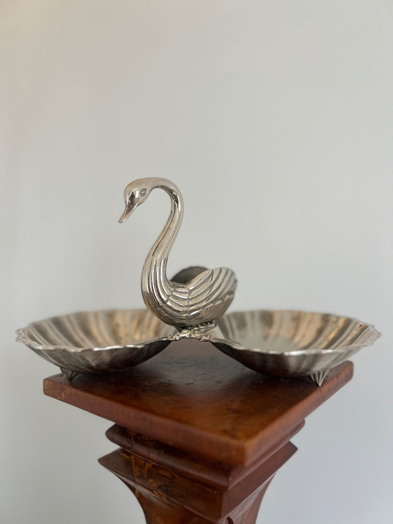 The Ornate Swan Shell Dish by Porter Studio is a decorative silver-plated swan figurine featuring two scalloped trays, elegantly displayed on a wooden pedestal against a neutral backdrop, merging art and functionality.