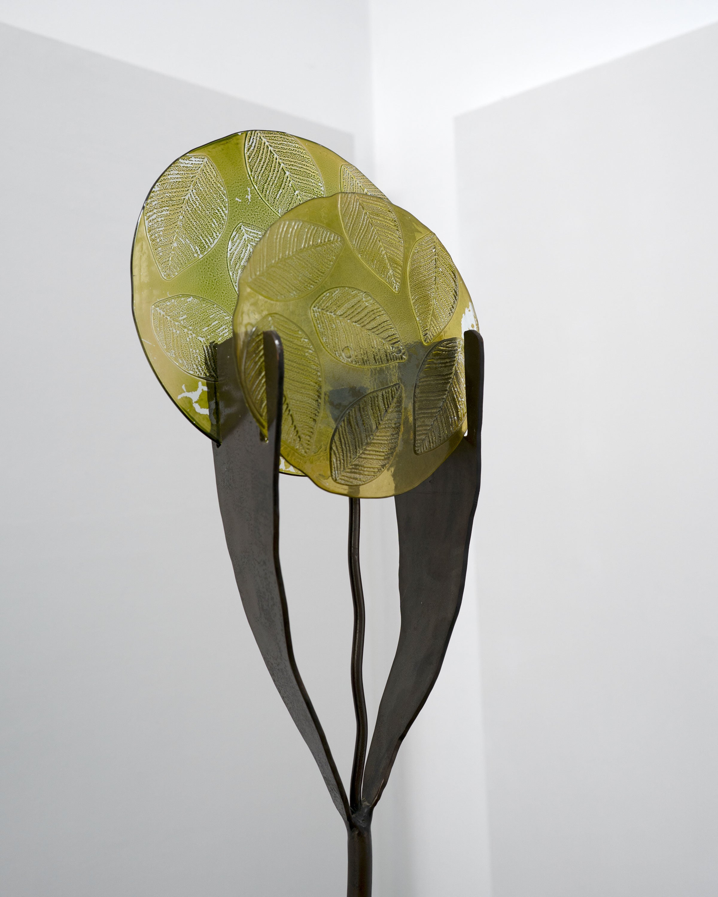 The Tom Bogle Brutalist Wrought Iron and Cast Glass Lamp (Norway, c.1970) features two yellow-green, leaf-patterned glass shades on dark metal stems and a wrought iron base, showcased in a minimalist, light-hued room.