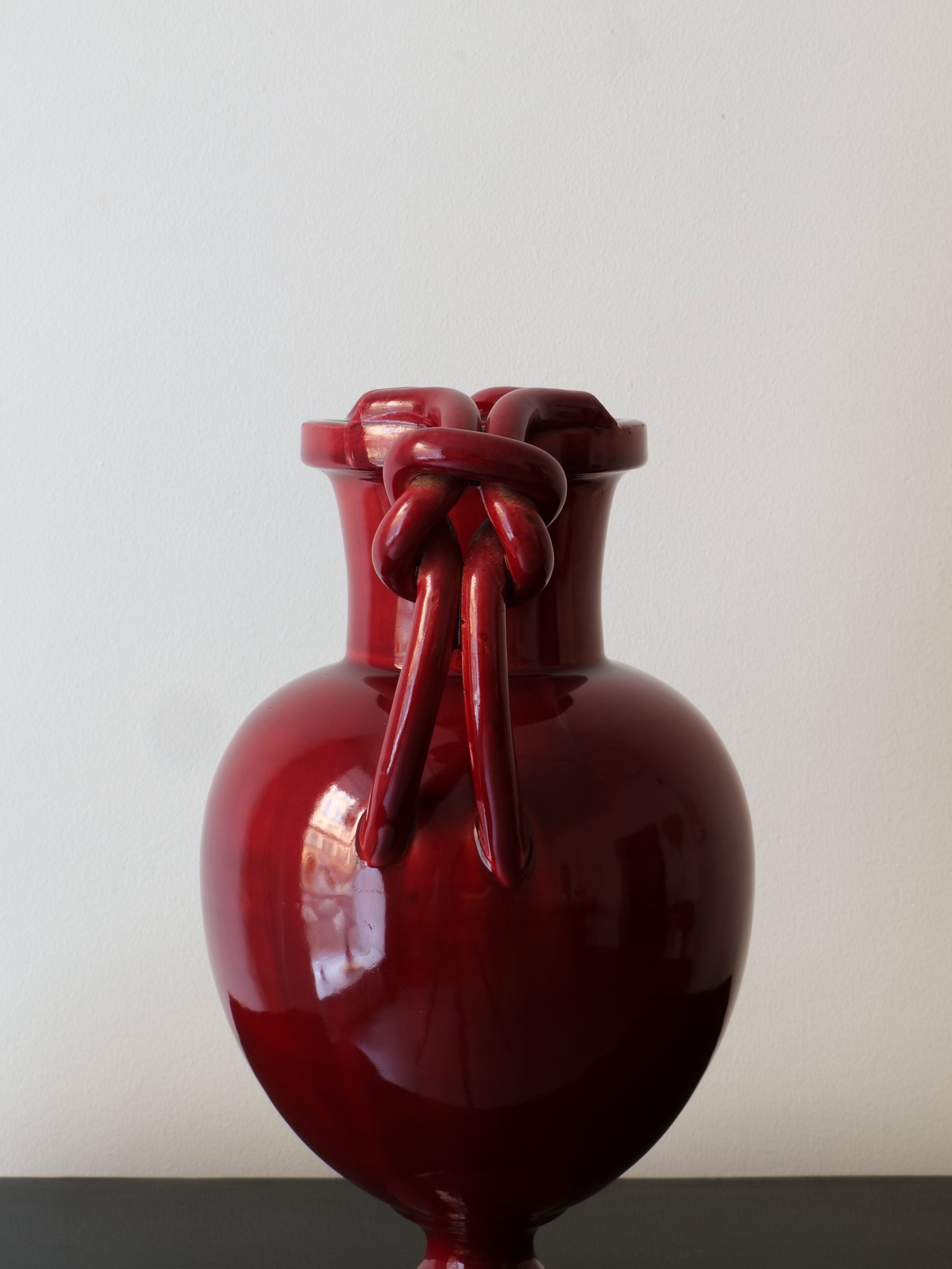 The Baluster Vase Jérôme Massier by Collection apart, with its deep red ceramic and glossy finish, rests on a dark surface exuding the charm of French Art Nouveau. Its top showcases intricate twisted handles reflecting the artistry characteristic of Vallauris ceramics, all set against a simple light-colored wall background.