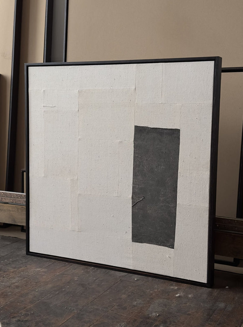 A framed, minimalist artwork featuring a textured white surface with rectangular patches of hand loomed tapestry and a single rectangular black patch on the right side. The artwork is leaned against a beige and brown background is the (a-tent), 2024 Framed Hand Loomed Tapestry by Bec Kirby.