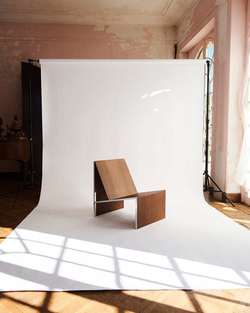 The Lounge Chair 2022 by Studio HAOS, crafted with minimalist design and simple materials, is displayed on a white backdrop in a sunlit room with pink walls and wooden floors, where sunlight creates dramatic shadows.