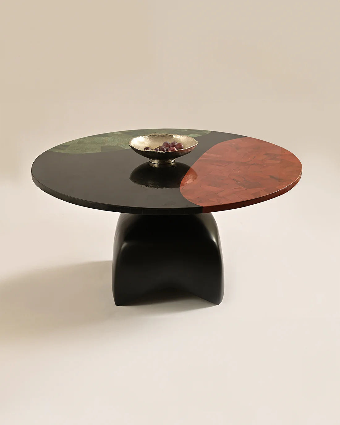 The LMNOH Jasper Coffee Table showcases a glossy, segmented top in black, green, and reddish-brown with semi-precious stones on a unique curved black casted aluminum base. A small silver bowl with grapes sits at its center against a plain off-white background.