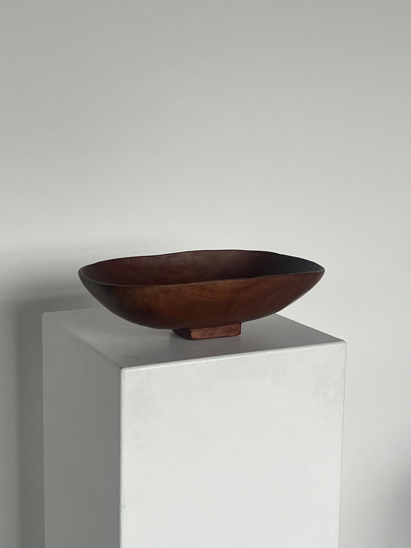 A dark, hand-carved Mahogany Hand Carved Oval Bowl from Porter Studio with a smooth finish sits on a tall white pedestal against a plain grey background. The oval piece, showcasing exceptional craftsmanship, features rounded edges and is slightly raised on a small base.