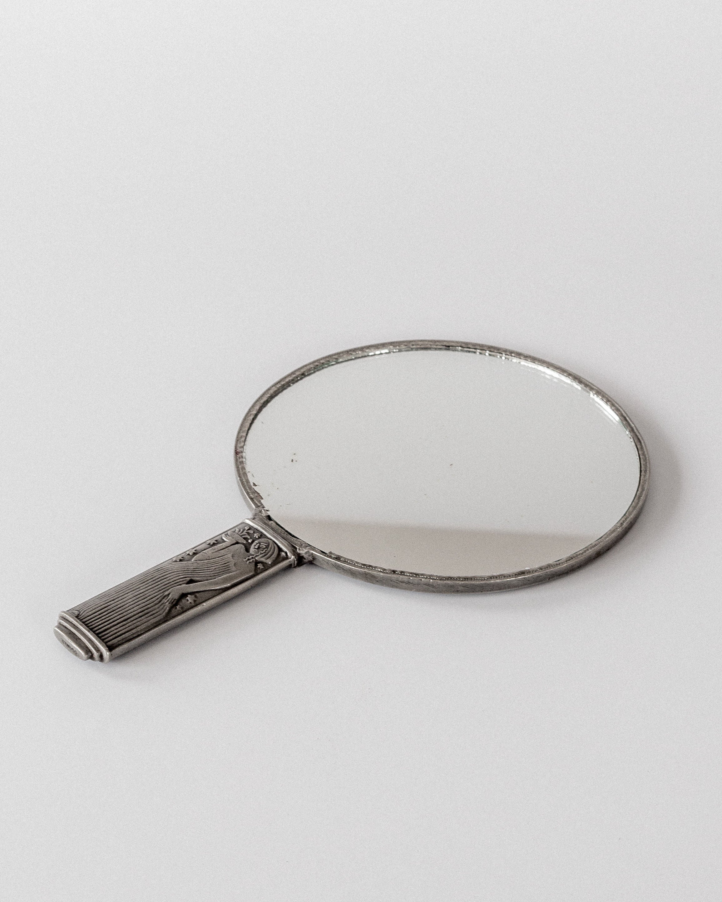 The Modernist Metal Mirror by Sylvia Stave, a 1930s design from Spigel, features a round mirror with an intricately engraved pewter handle. It rests on a plain white surface, slightly tilted to reflect the stark background, echoing timeless elegance.