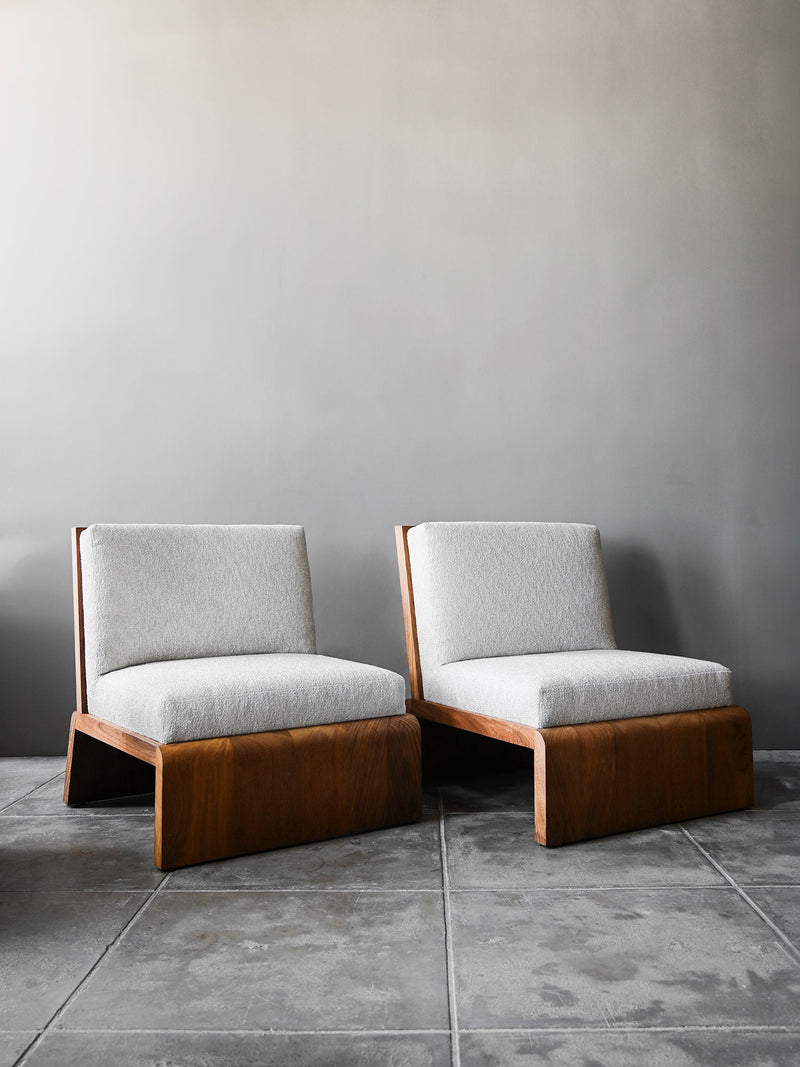 Two Saint Maxine Outdoor Chairs by Studio OSKLO, featuring teak frames and light gray cushions, sit on a tiled floor against a plain gray wall, creating a minimalist and contemporary atmosphere.