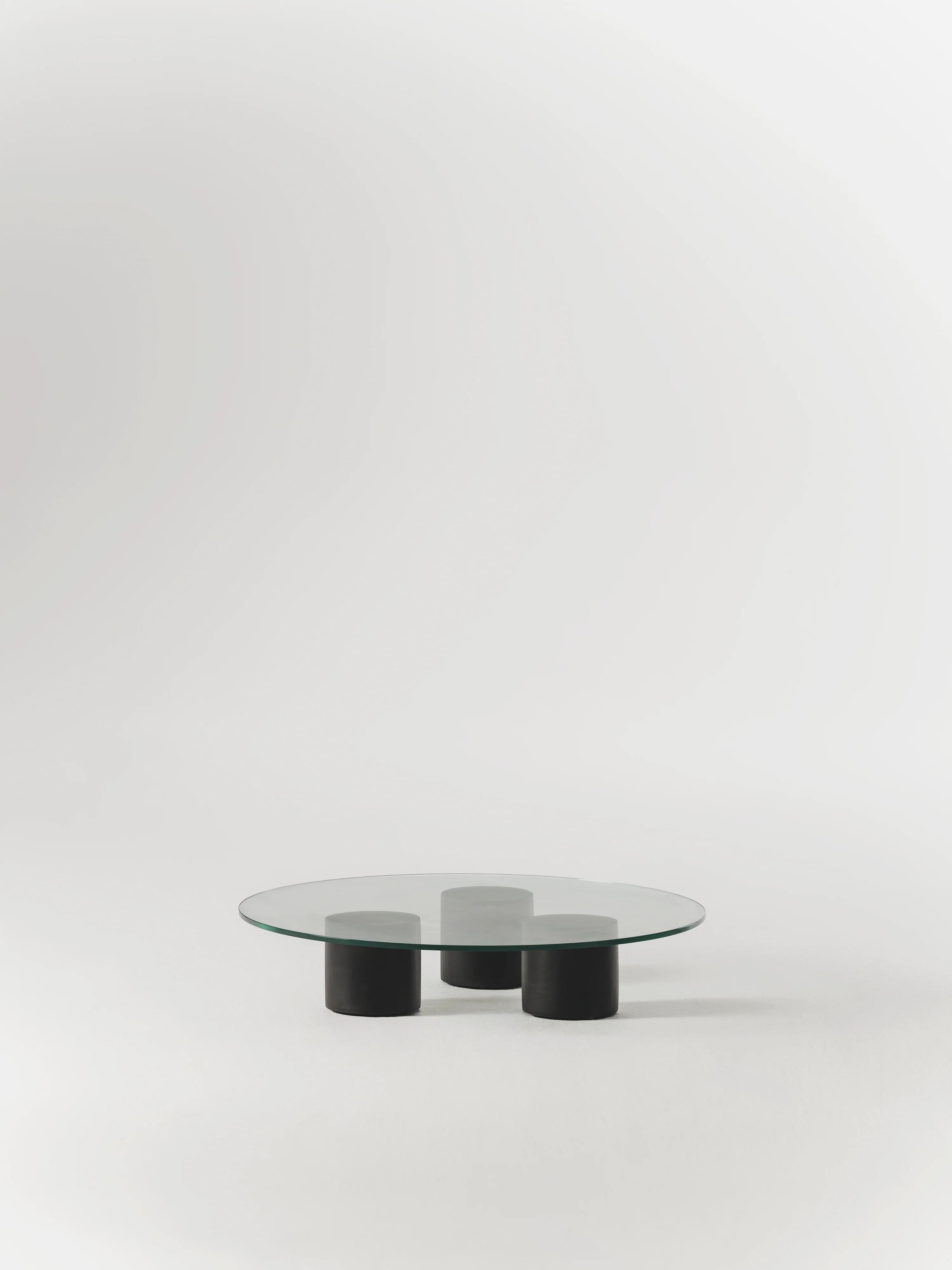 The Carmene Glass Coffee Table by MarlotBaus showcases a circular tempered glass top with three cylindrical black legs, set against a plain white background.