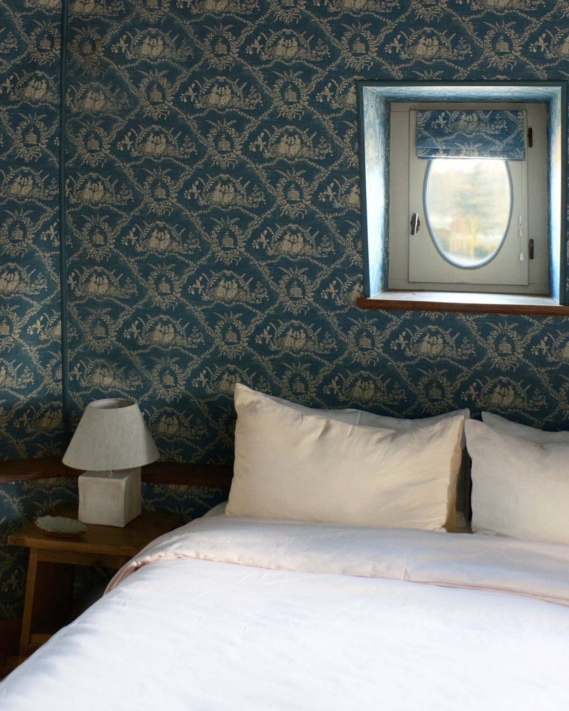A cozy bedroom features a bed with white linens, a nightstand topped with the Project 213A Arouca Table Light White, and blue floral wallpaper. A small window with a blue-patterned shade fills the space with natural light.