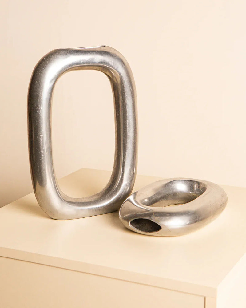 A pair of Italian aluminum vases from the '80s by Treaptyque rest on a light-colored surface. One is tall and elongated, resembling a vertical loop, while the other is shorter and wider with a circular opening. Both exude Italian elegance with their shiny, reflective finish.