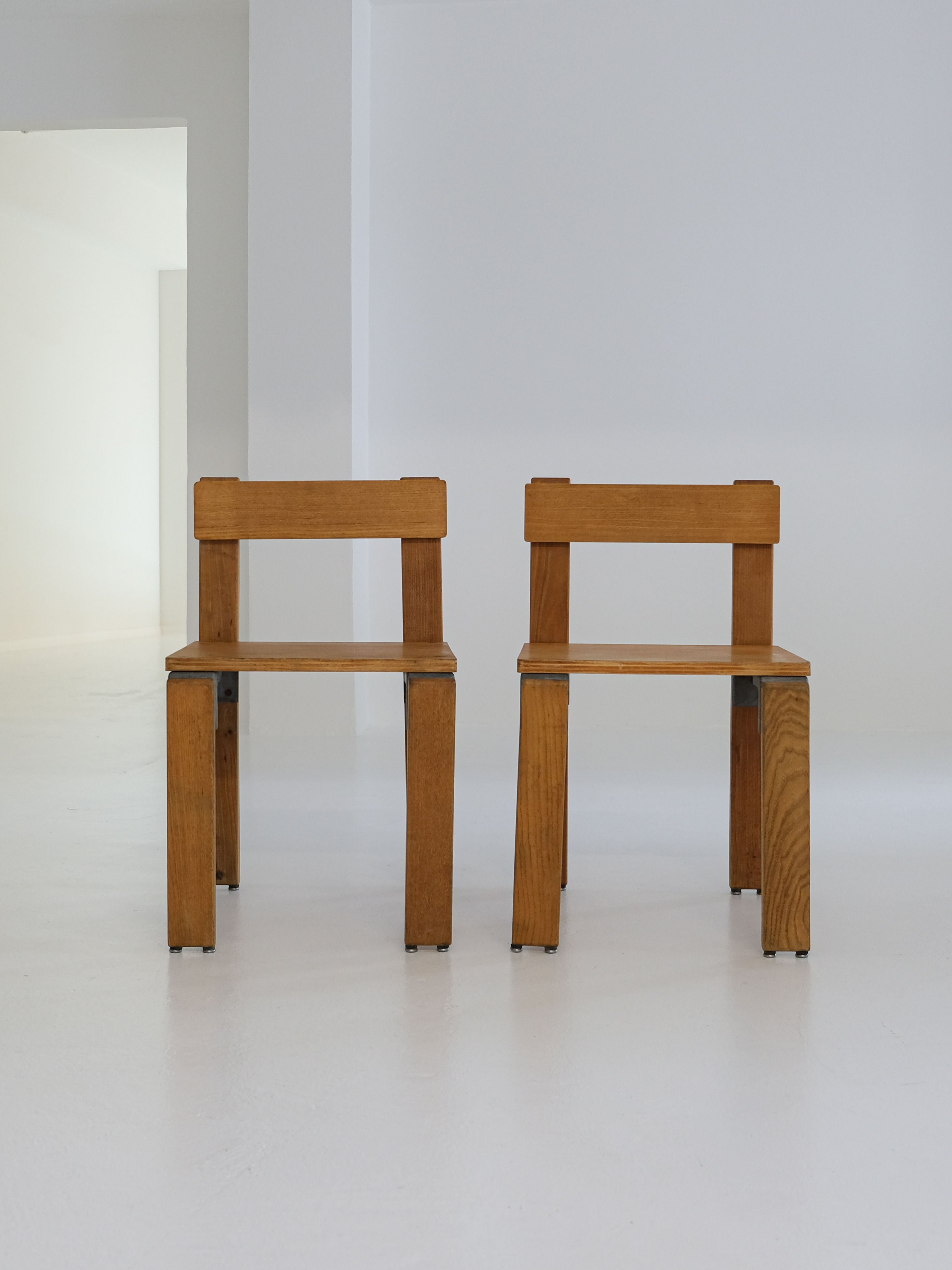 The minimalist room features two "Les Carats" dining chairs by George Candilis and Anja Blomstedt from the 1970s, crafted from oak plywood with simple geometric designs. Their natural wood texture contrasts beautifully against the smooth, white floor by antibeige.