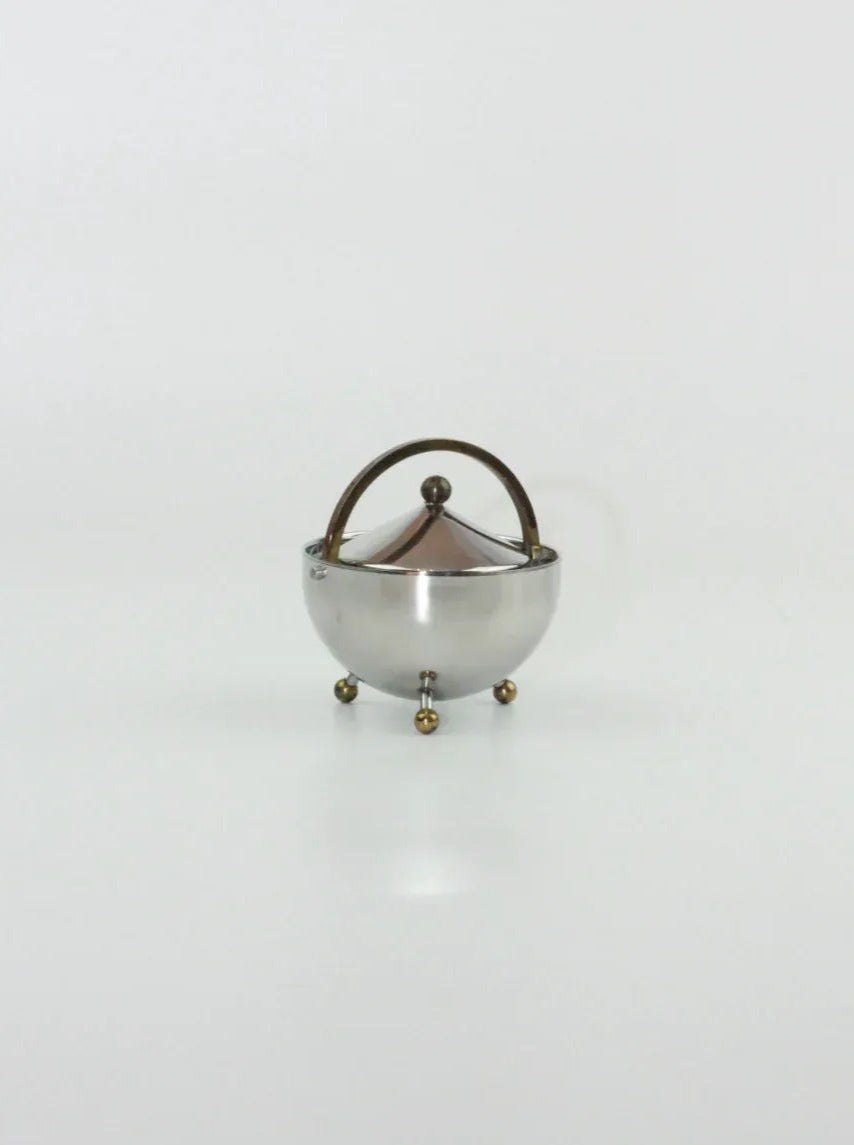 A small, round, metallic container on three ball-like feet sits against a plain white background. Designed by the renowned Danish designer Carsten Jørgensen for Boga Avante Shop, it features a curved handle and a pointy, conical lid. Crafted from stainless steel with brass accents, the Danish Sugar and Creamer Set exudes modern elegance.