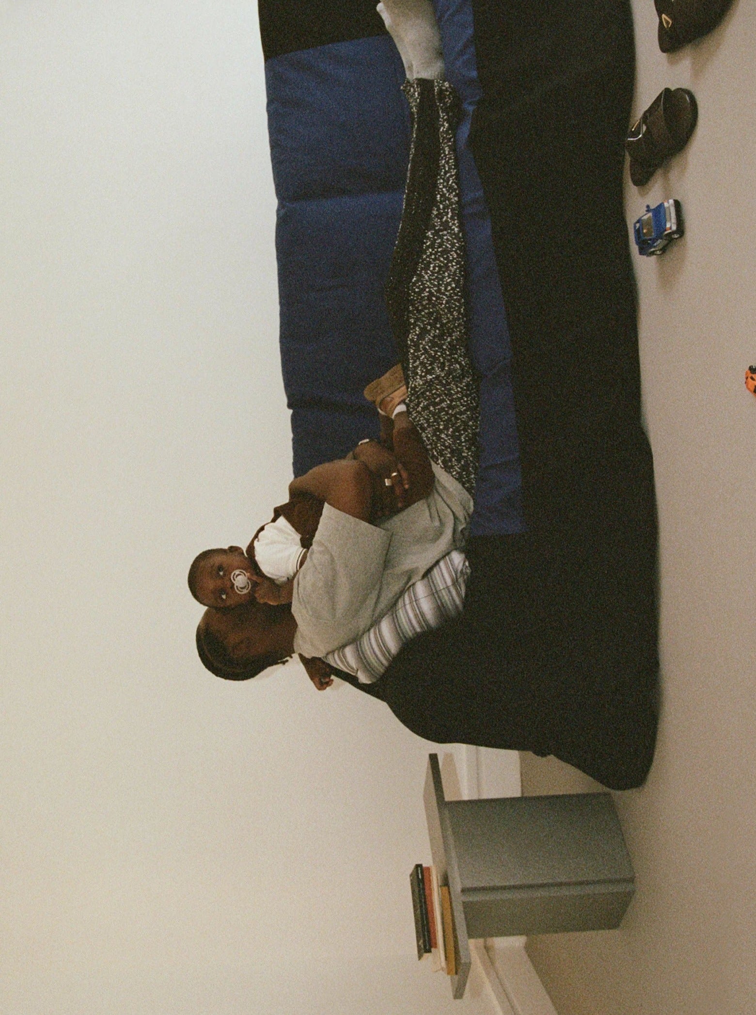 A woman sitting on the floor lovingly holds a baby with a pacifier. The baby, dressed in white, is close to a bed adorned with a blue and black blanket. A "Mara T" Side Table – Silver Noise by TEGET complements the scene, featuring laminated furniture and a few small toys nearby.