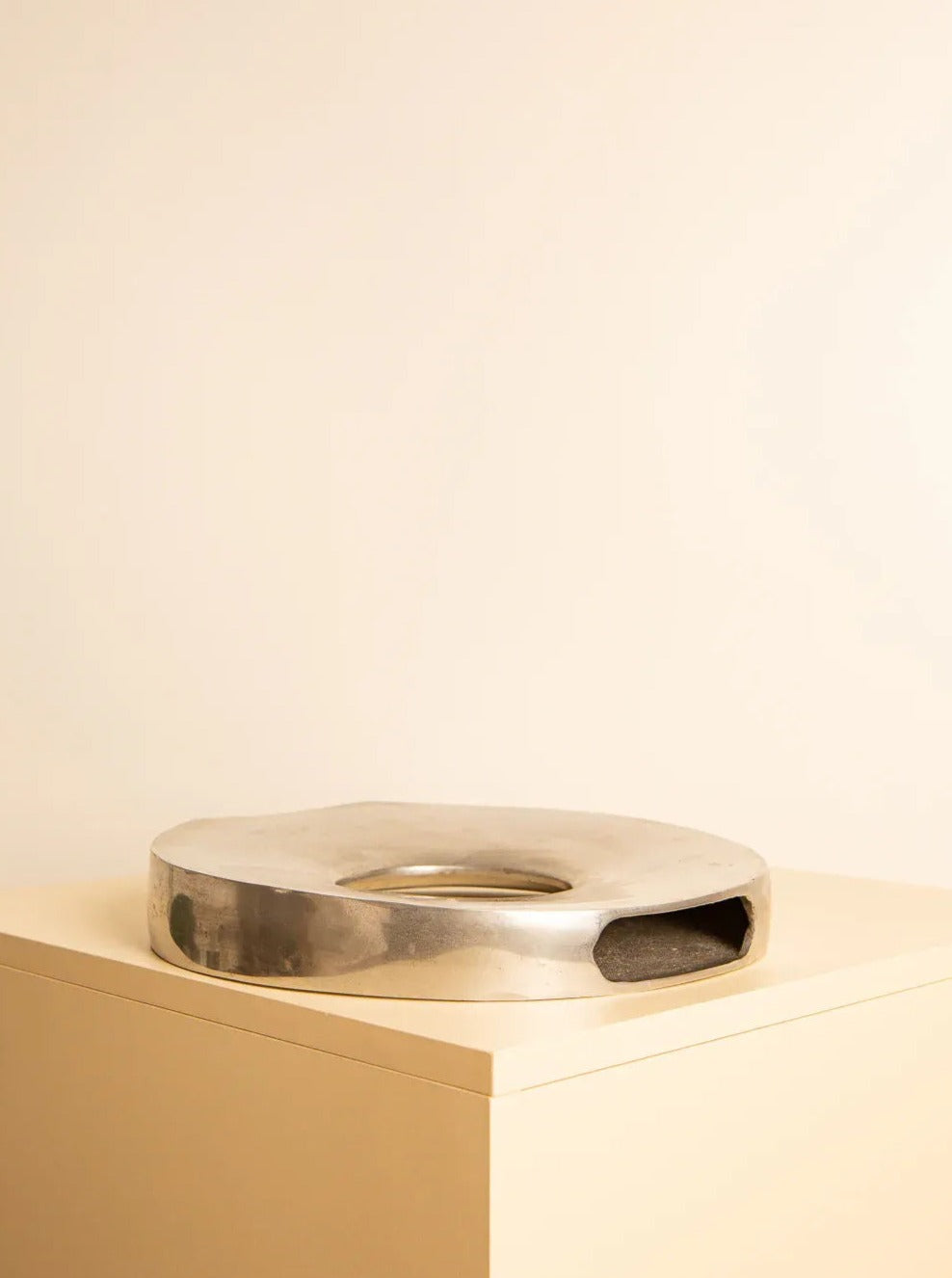 A polished, circular silver sculpture with abstract cut-outs sits on a flat, light-colored pedestal against a neutral background. The contemporary doughnut shape of this piece showcases Italian craftsmanship, reflecting light beautifully on its smooth surfaces. The "Donut" Vase in Aluminium 80's by Treaptyque exemplifies modern design with a touch of elegance.