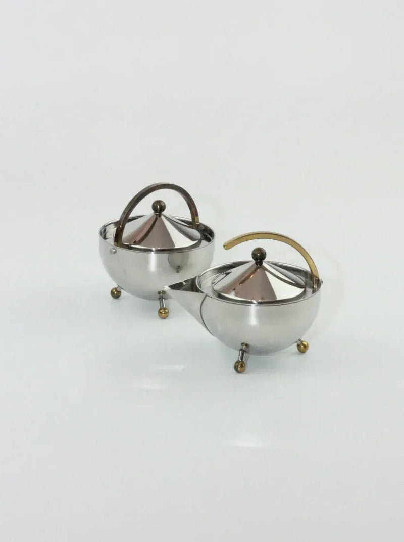 A pair of spherical stainless steel Danish Sugar and Creamer Set from Boga Avante Shop, embodying Danish design, with brass handles and finials. Each piece stands on three brass and steel ball feet and has a lid with a conical shape. The background is plain white.