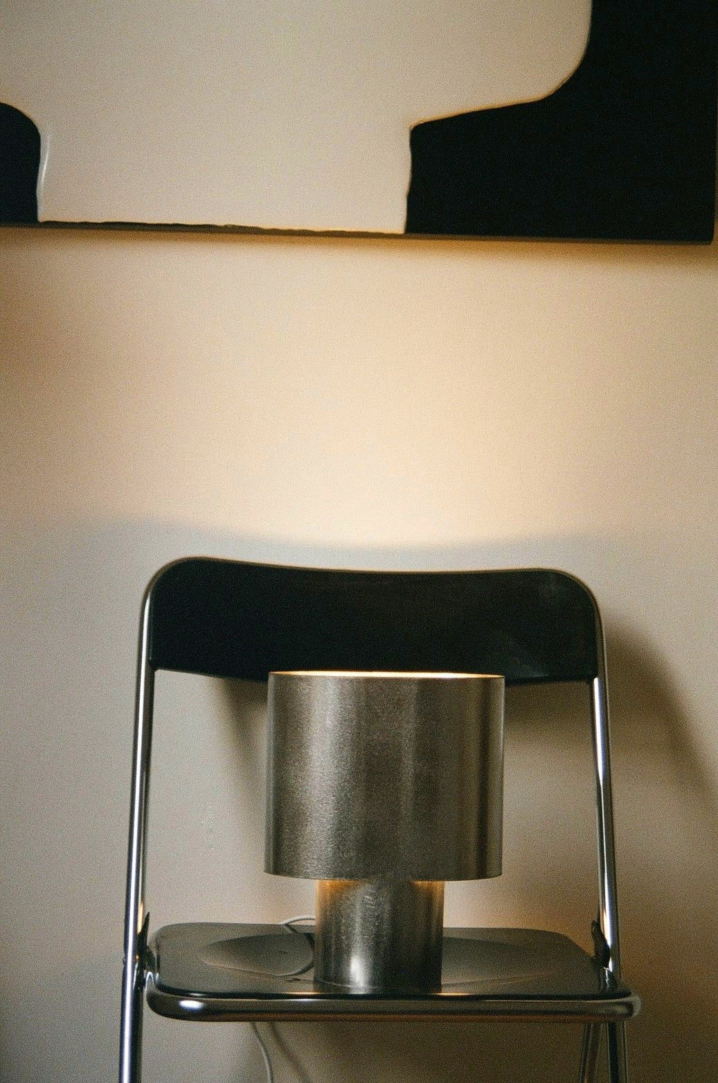 A Bien Paris Table Lamp in Brushed Steel rests on a black folding chair, set against a beige wall with a modern abstract black and white painting overhead, creating a timeless design scene.