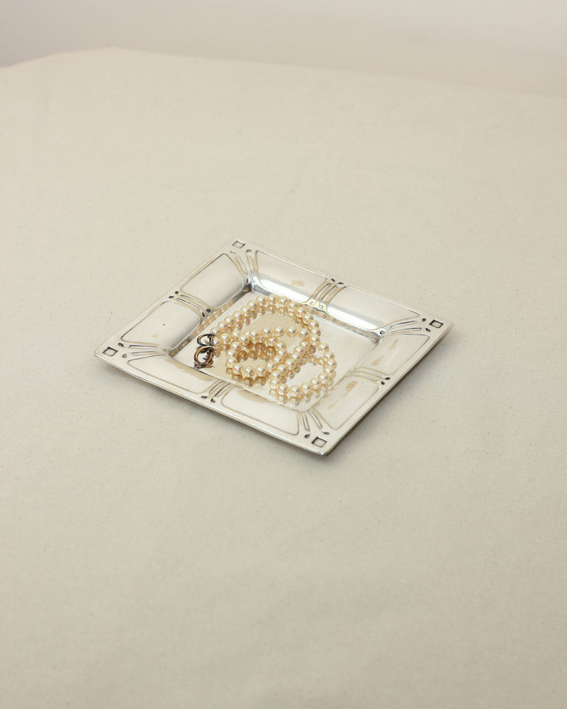 A Silver Modernist Vide-poches by Boga Avante Shop, featuring a decorative pattern, holds a pearl necklace on a light beige surface with a minimalistic background.