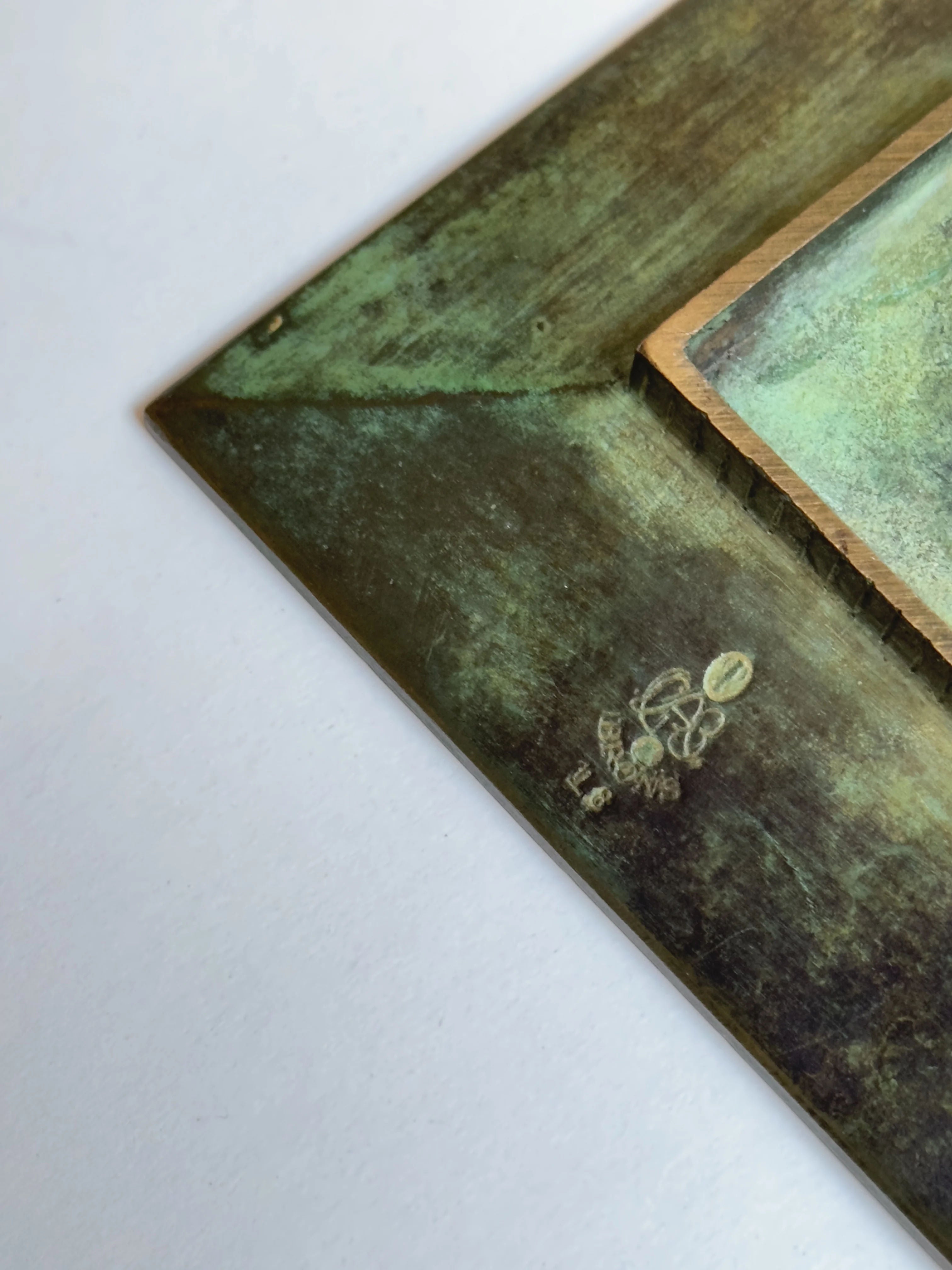 Close-up of a textured artwork corner with a metallic aged green and bronze look, featuring a small embossed emblem and serial number. It has an antique or industrial vibe, similar to Galerie Storm's GAB Bronze Dish with its rich patina, reminiscent of Jacob Ängman's style.