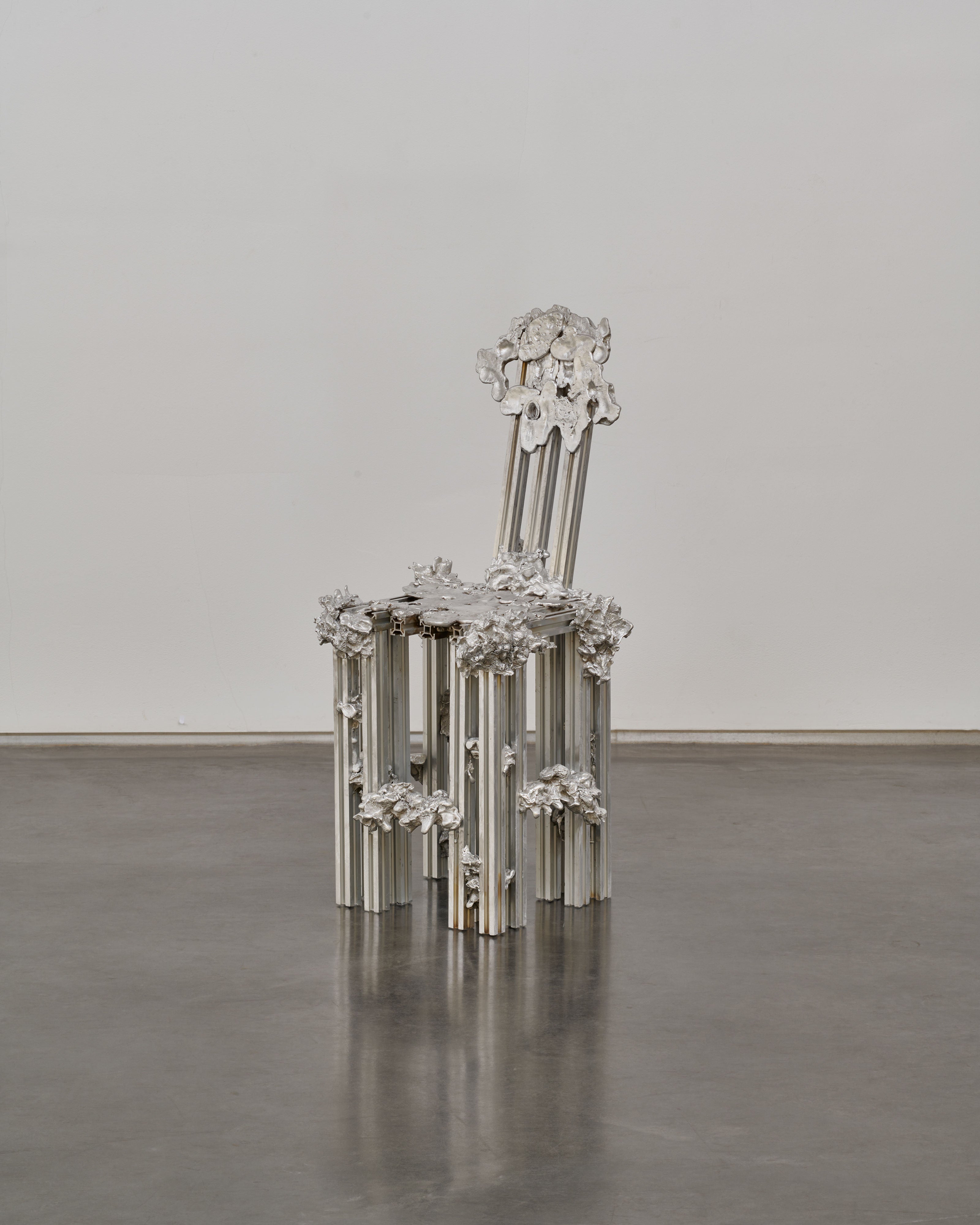 The 'Graft' Chair in Cast Aluminum by Sheyang Li is a silver chair with an ornate, sculptural design featuring flower-like details and vertical rods, set against a white wall and polished gray floor.