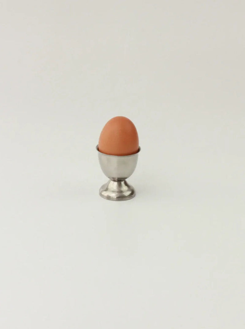 A single brown egg is placed upright in a small, elegant Boga Avante Shop Set of Six Stainless Steel Egg Cups against a plain white background.