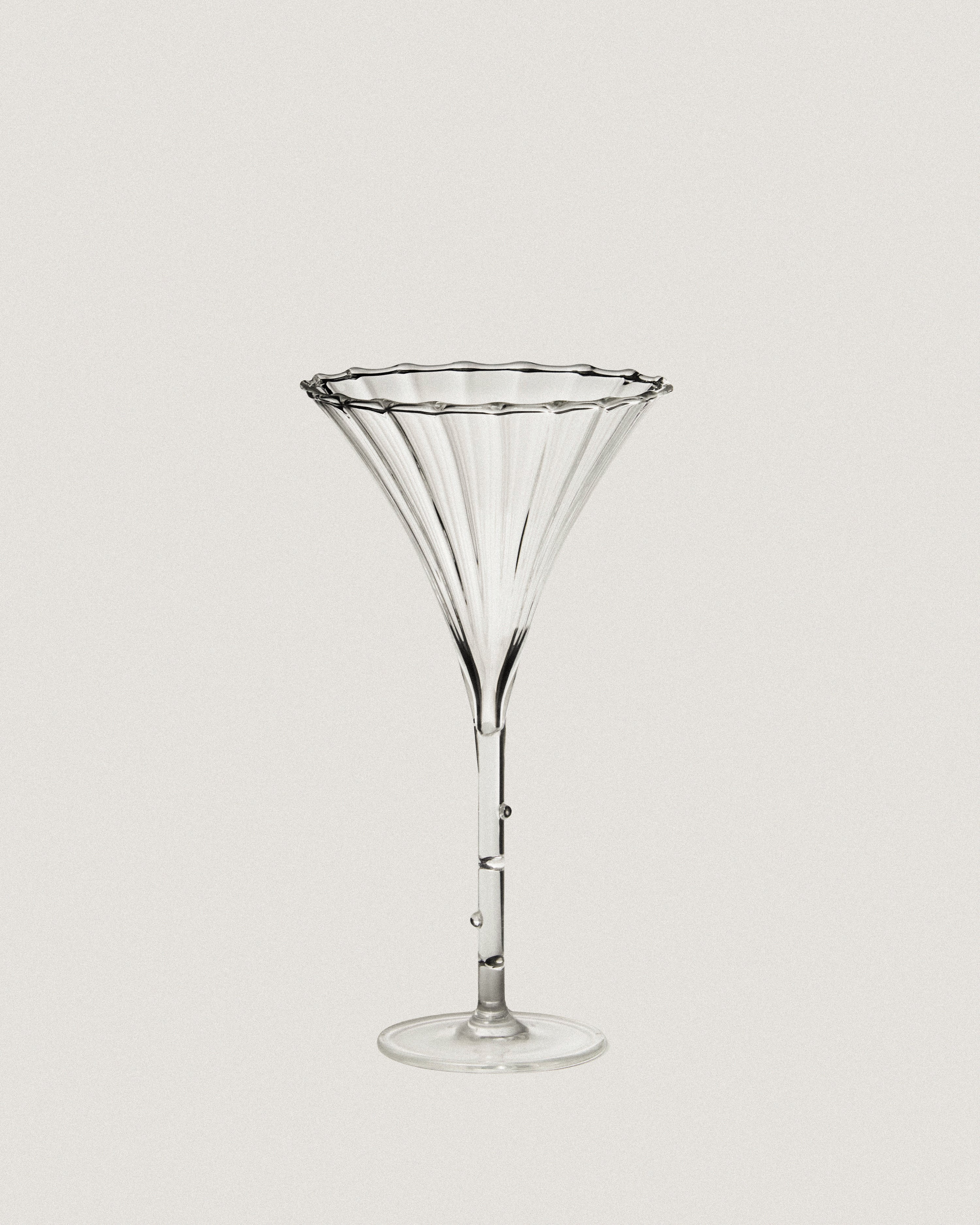 A clear, fluted Trumpet Aperitif Glass by Sophie Lou Jacobsen with a wide, wavy rim sits on a tall, slender stem and circular base, all set against a plain white background. The elegant design features subtle vertical ridges and a small bead-like accent on the stem. This aperitif glass is part of a stunning set of 4.