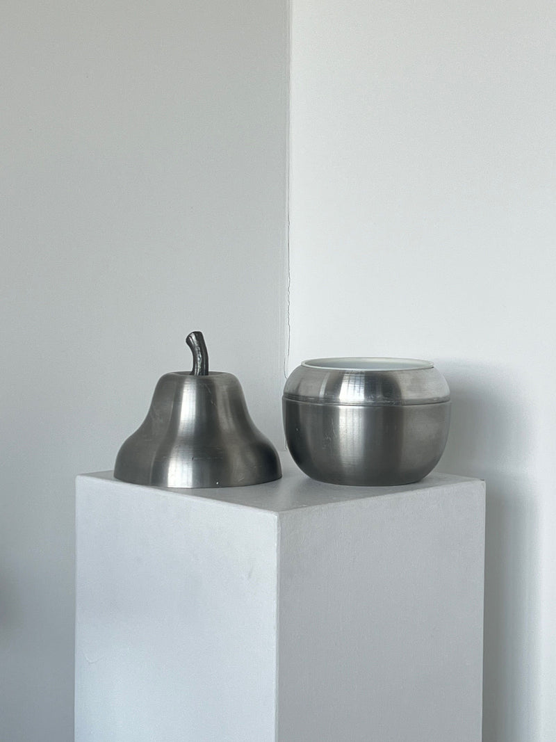 A minimalist photograph features two metallic objects on a white pedestal. On the left, a Porter Studio Pear Wine Cooler in brushed aluminium with a stem stands prominently. On the right, there is a round, smooth bowl of brushed aluminium. Both pieces are striking against the simple white background.