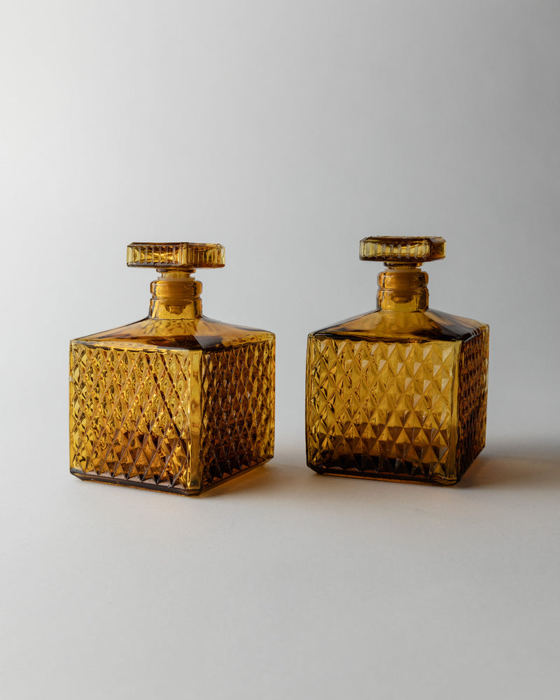 Two Bottega Jacobs Amber Glass Decanter 70s, featuring square shapes, vintage diamond point patterns, and flat round stoppers, are placed side by side on a plain, light background.