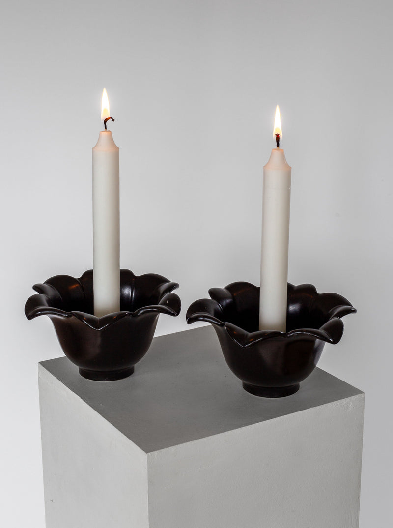 Two lit white candles stand in dark, flower-shaped holders made of ceramic. These Mid-Century Ceramic Candlesticks by Spigel from 1950s Sweden are placed side by side on a minimalist gray pedestal, set against a plain white background. The soft glow of the candle flames illuminates the scene.