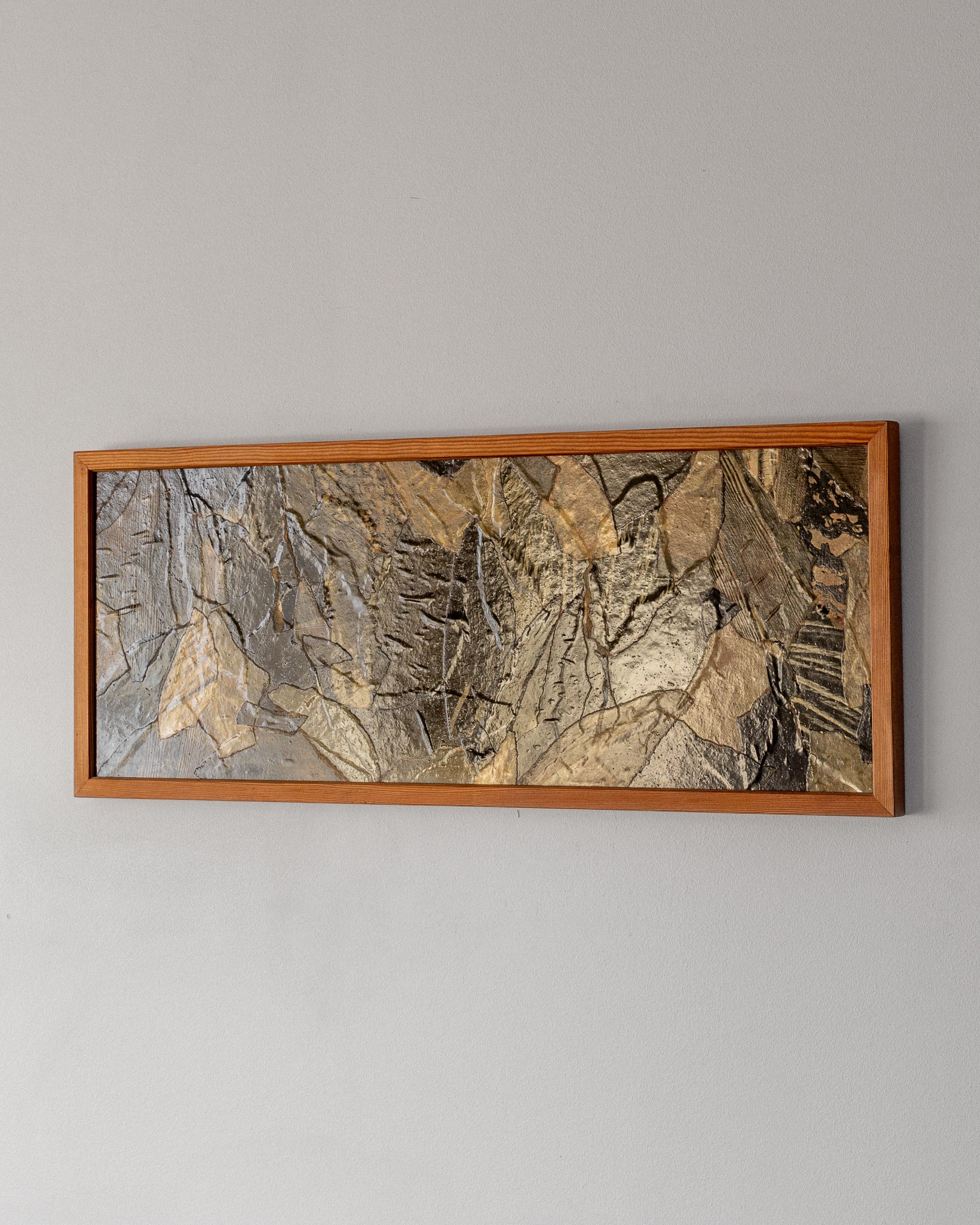 The artwork "Leaves" by Lars Bergander, branded by Spigel, hangs on a gray wall with a wooden frame. It captures the mid-century modern Scandinavian style, featuring an abstract collage of overlapping stone-like pieces in gray, brown, and beige for a textured look.