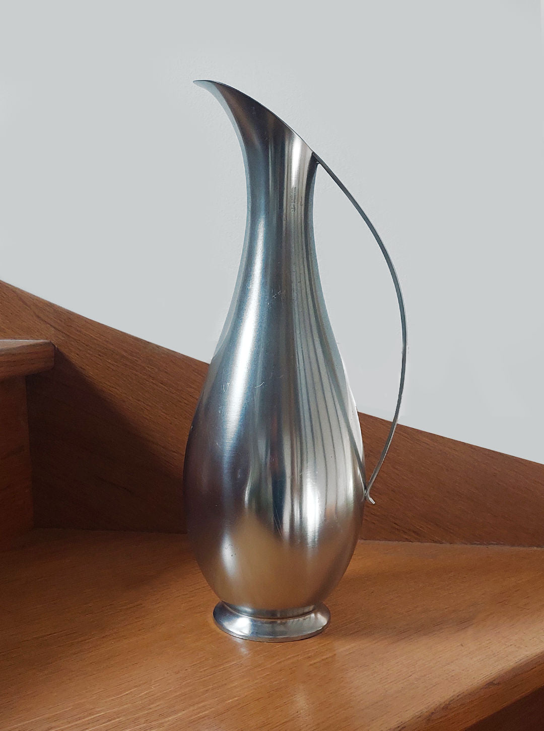 A sleek, silver jug from Les Objoies, labeled as the Large Pewter Carafe, with a narrow spout and elegant handle, graces a wooden staircase. The carafe's contemporary design showcases a modern, reflective surface and curvy lines that stand out against the neutral backdrop.