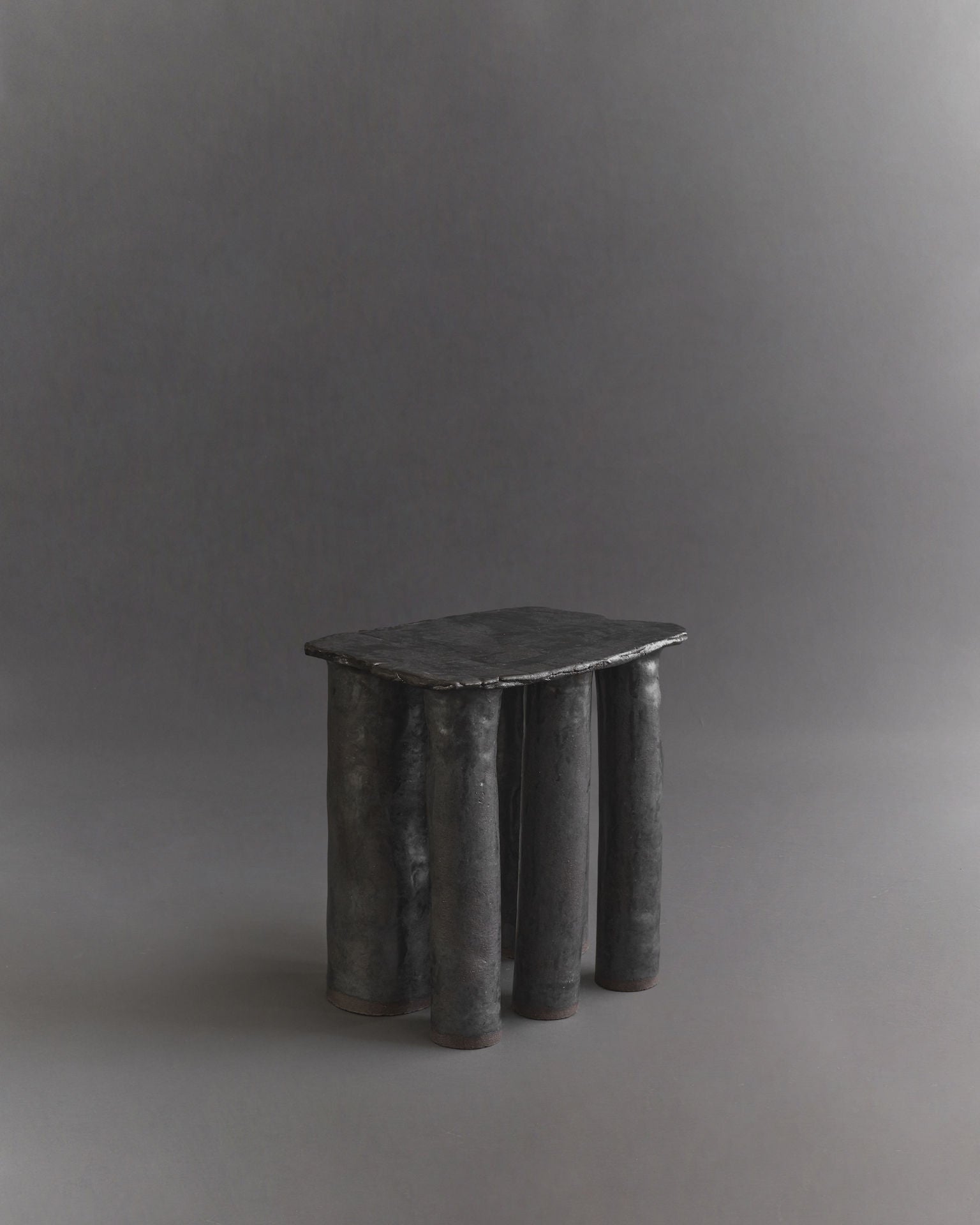 The Cleo Ceramic Side Table by Ombia Studio is a small, handmade sculptural table featuring an irregular oval top and cylindrical legs with a textured black glaze, set against a plain gray background.