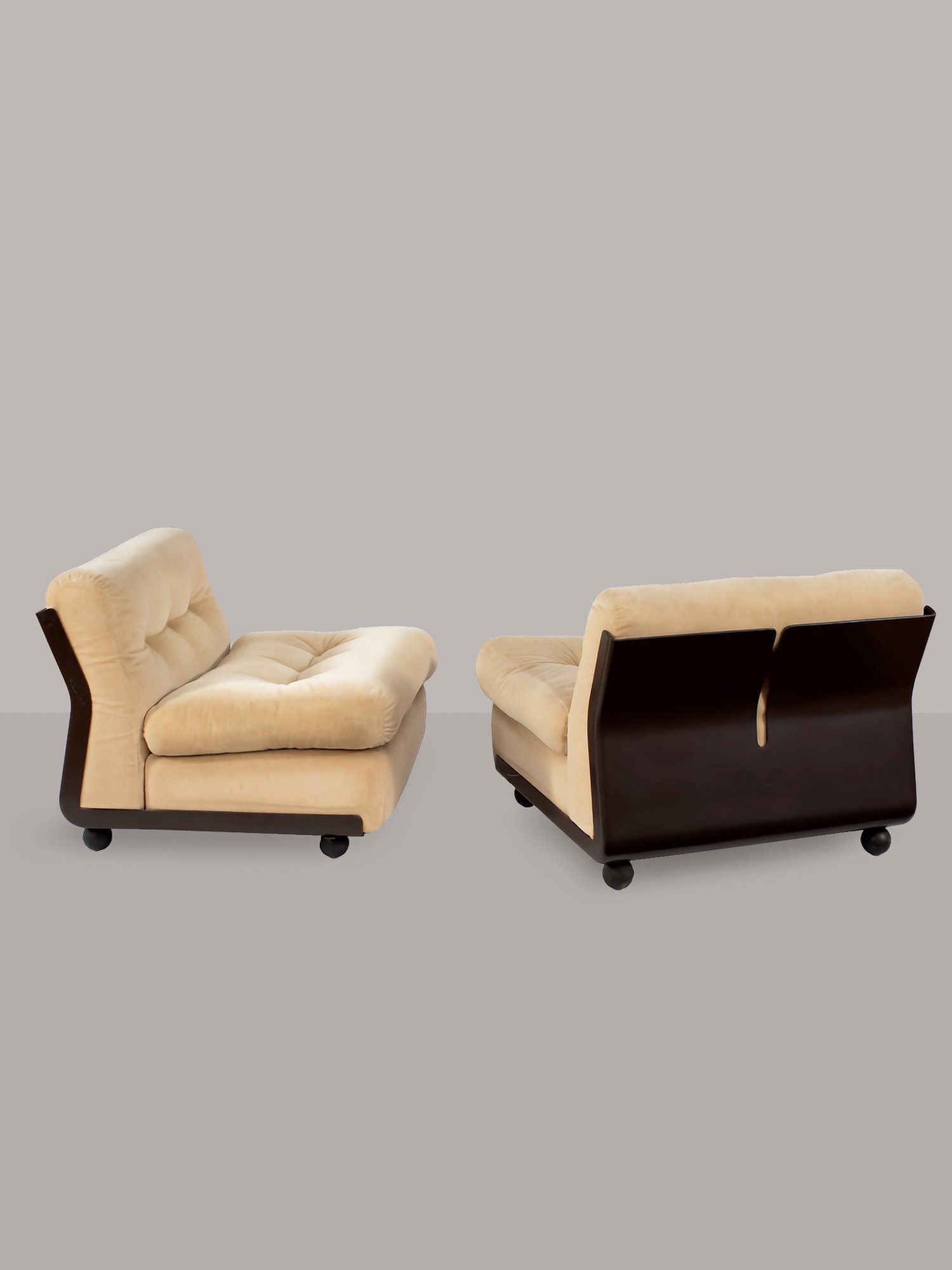 Two Amanta Armchairs designed by Mario Bellini in 1966 for RELIC LONDON feature beige cushions and dark wooden frames. Each chair has a low, rounded base with wheels, angled backrests, and a minimalist design reminiscent of the iconic Amanta armchair. Set against a plain grey background, these chairs perfectly showcase their modern aesthetics.