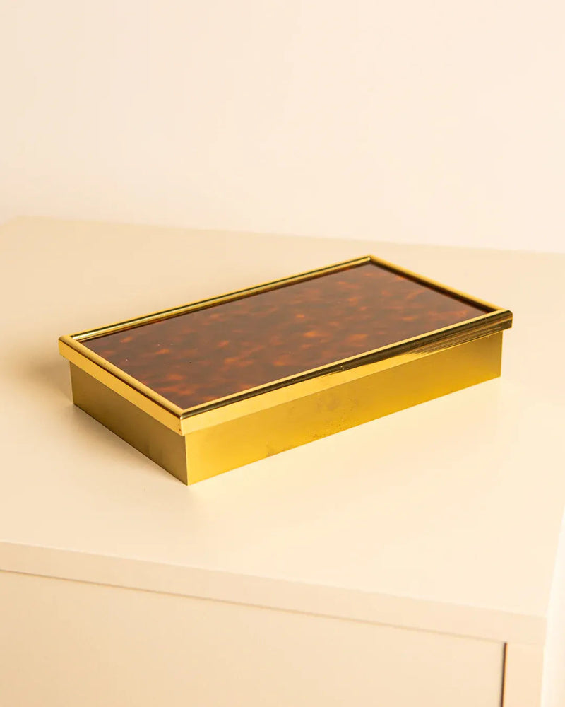A rectangular, gold-colored Italian Box by Bicchielli 80's from Treaptyque sits on a cream table. Its reflective finish and tortoiseshell-patterned lid echo classic elegance, embodying a simple yet modern aesthetic.