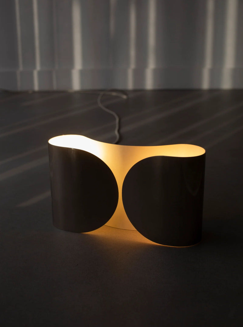 A modern floor lamp with an abstract, dual-panel design emits a warm, ambient glow reminiscent of the Chrome Wall Lamp by Tobia Scarpa 80's from CU-RA-TED. The black panels radiate a soft light from within, casting patterned illumination on the nearby wall as it sits on a dark floor with its visible power cord extending away.