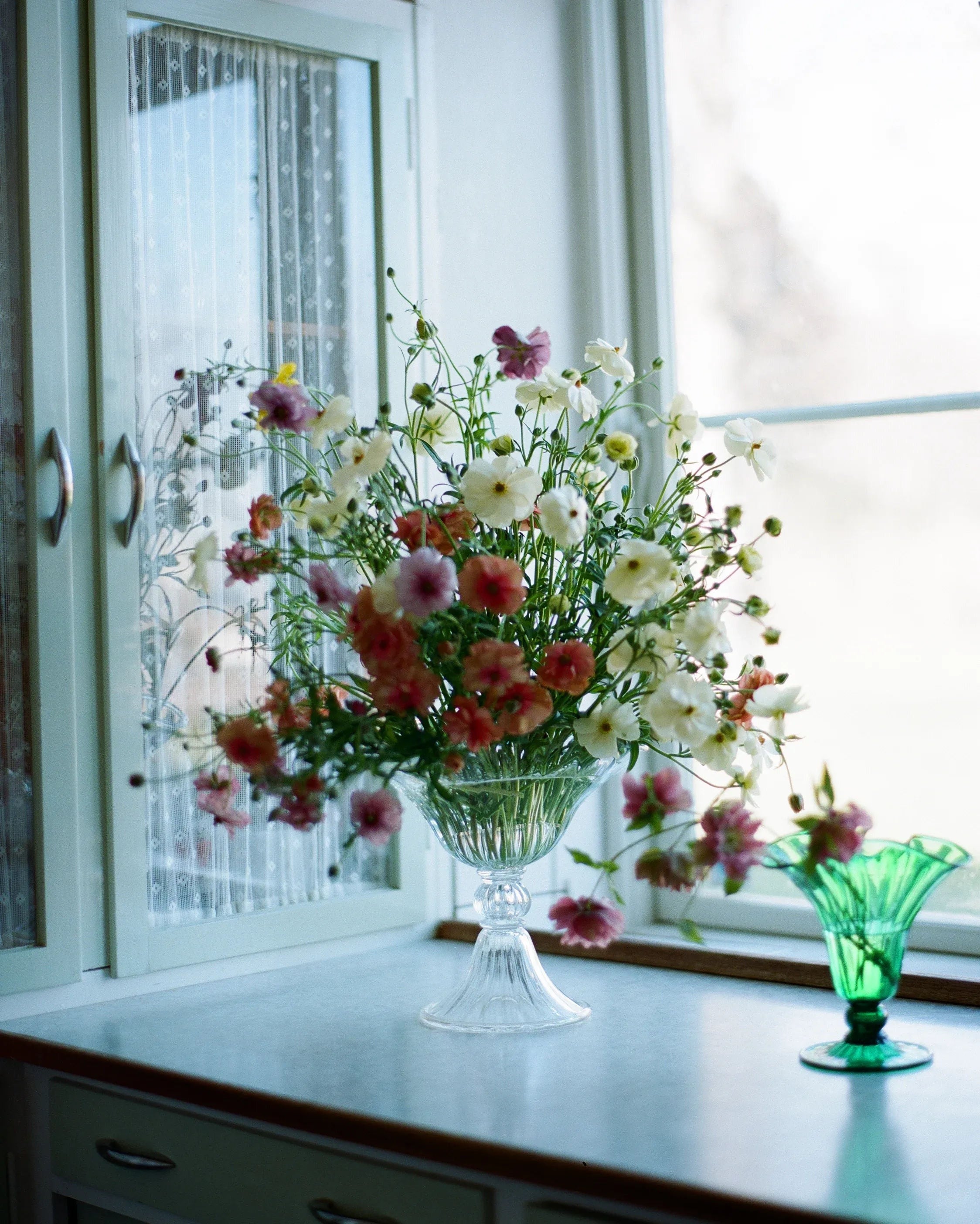 A large pedestal vase brimming with pink and white flowers is set on a white countertop next to a window. Beside it is an Akua Objects Karen Blixen Vase Small. The background features a cabinet with decorative glass doors. Natural light streams in, creating a bright and airy ambiance.