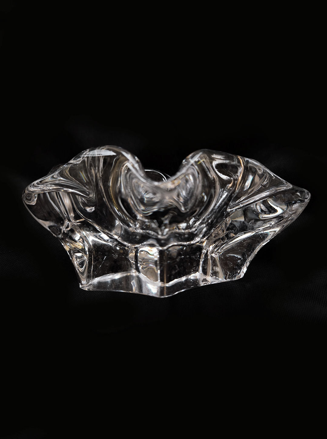 The "Glass Organic Ashtray" by Les Objoies features a clear, intricately designed glass with smooth, curved shapes resembling an artistic bowl. Set against a solid black background, it reflects light for shimmering highlights and a sculptural appearance.