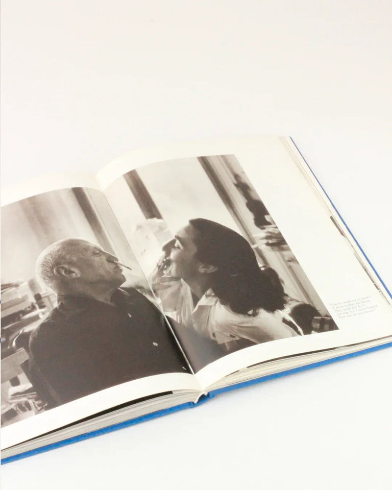 An open book displaying a black and white photograph of Picasso and Jacqueline sitting closely together, looking at each other in an intimate portrait. The person on the left is older and the person on the right appears to be younger. Picasso and Jacqueline from Boga Avante Shop is resting on a flat, light-colored surface.