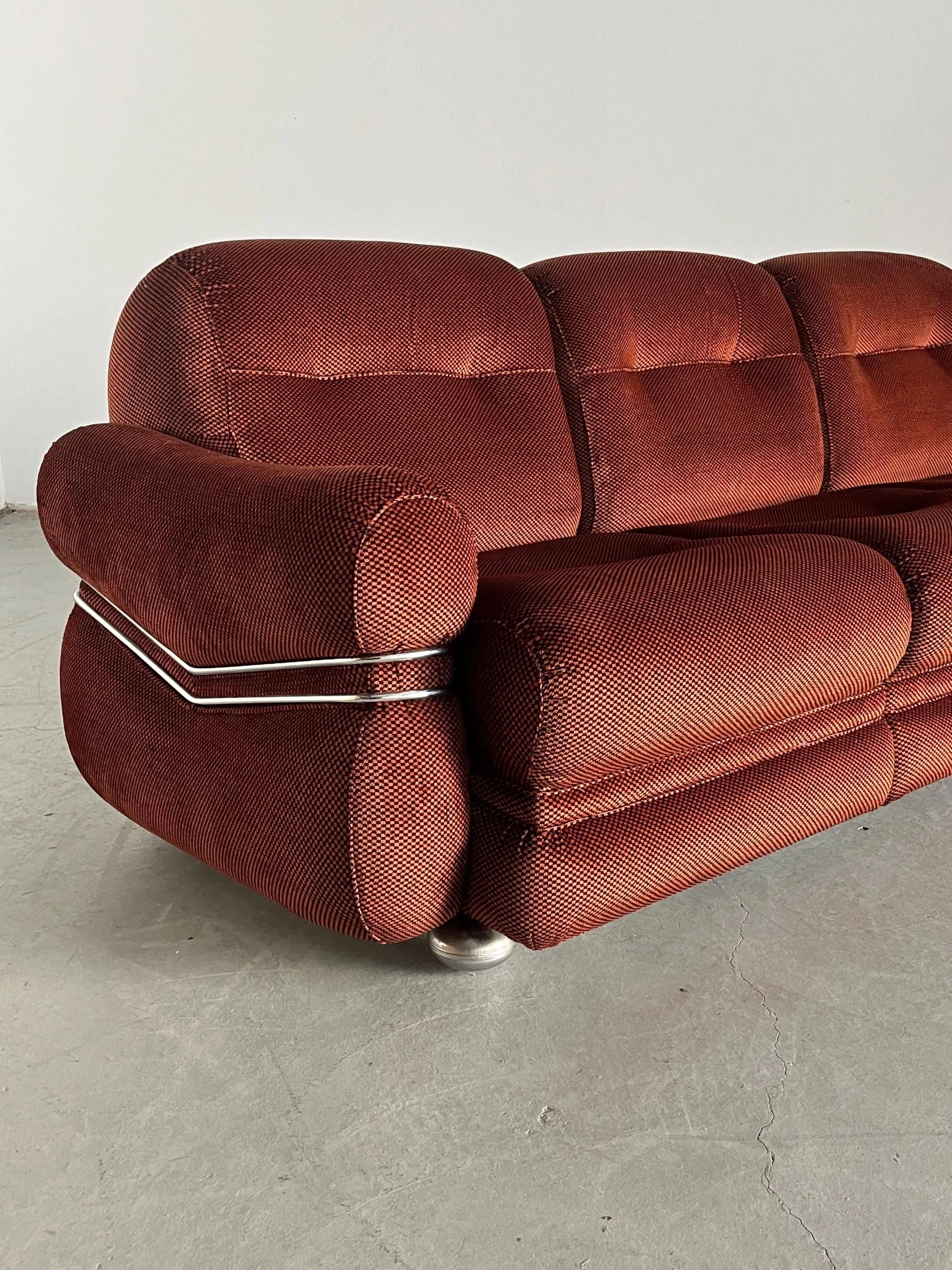 A Cherry Cargo Italian Space Age Seating Set from the 1970s features checkered upholstery, plush cushions, and rounded armrests. This vintage sofa exhibits mid-century modern design with metal accents, resting on small round metal legs against a plain light wall on a bare concrete floor.