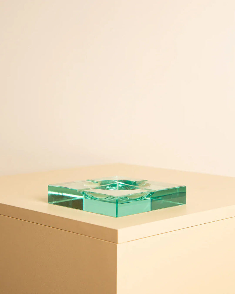 A clear, square glass ashtray with a greenish tint sits atop a simple, beige pedestal against a matching beige background. This Glass ashtray by Fontana Arte 70's from Treaptyque boasts a modern, minimalist design, epitomizing elegant Italian design and adding sophistication to the scene.