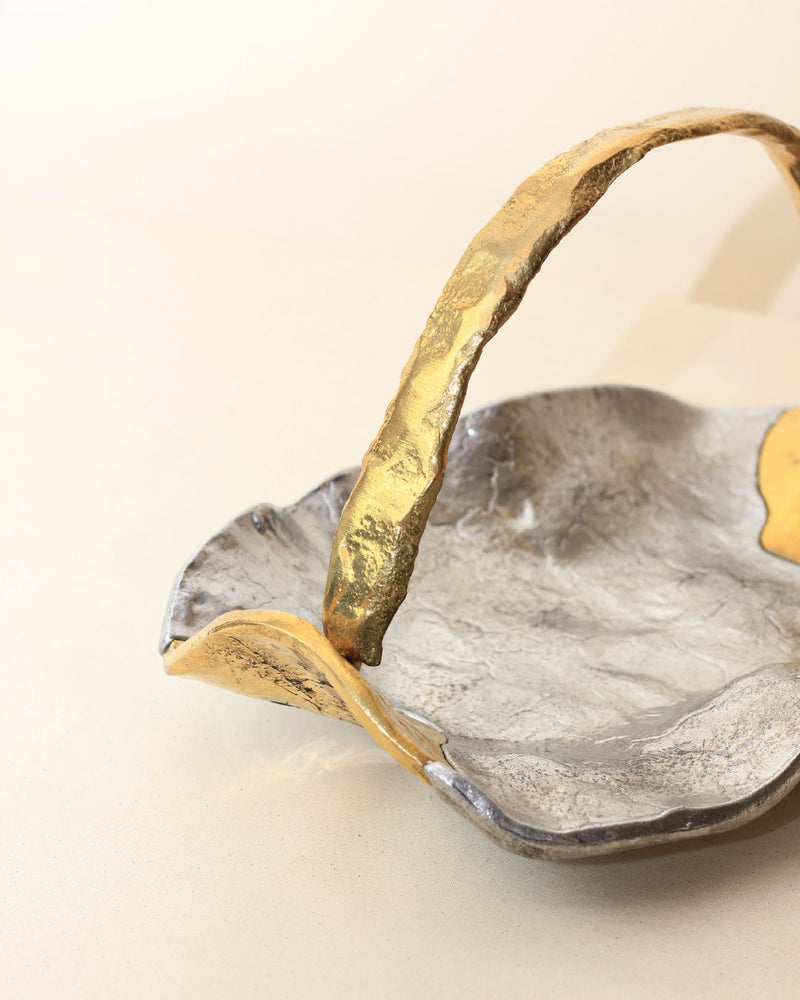 A close-up of the Brutalist Decorative Bowl by Boga Avante Shop—this silver dish has an irregular, wavy shape, a rustic finish, rough gold-toned textured accents, plus a curved handle and distinctive edge detailing.