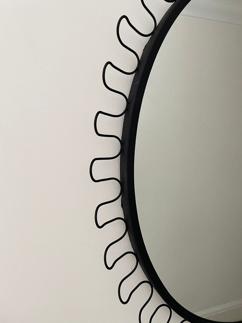 The Maud Vaughan Post Modern Oval Mirror, featuring a black steel frame and wavy sun ray accents inspired by mid-century design, hangs on a plain white wall. Only half of the striking mirror is shown in the image.