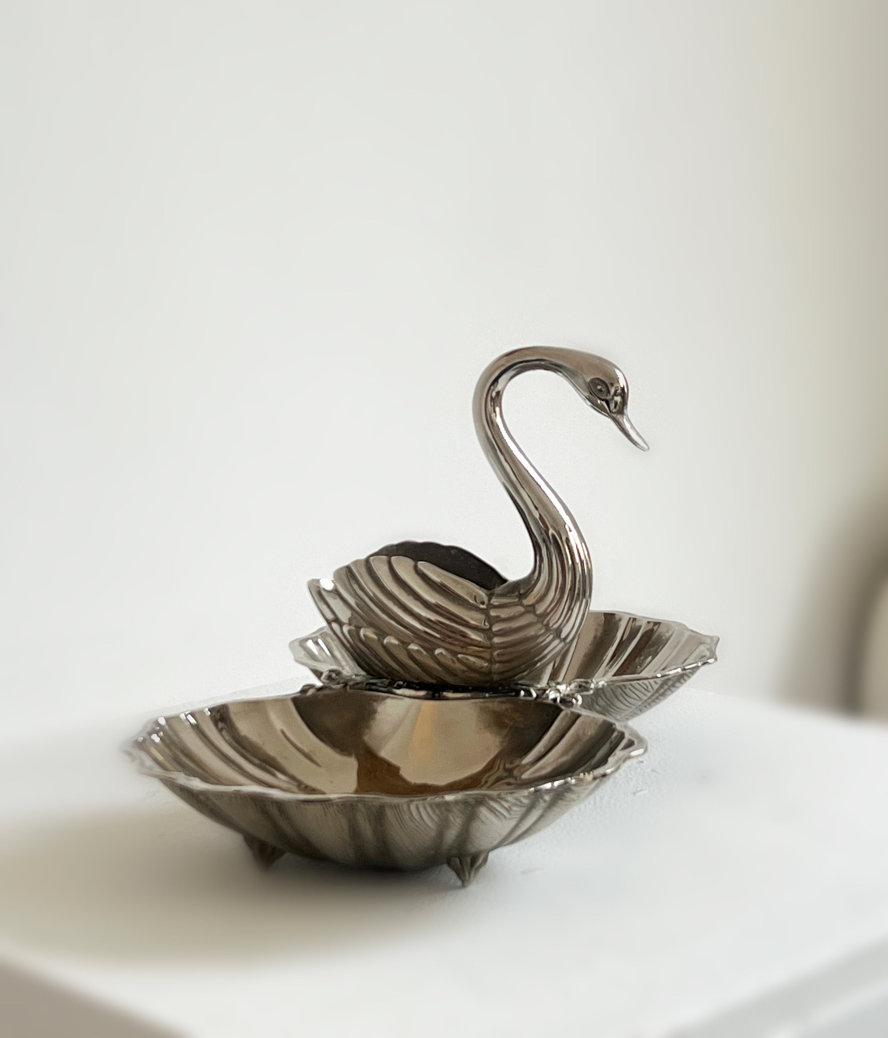 The Ornate Swan Shell Dish by Porter Studio features a silver-plated swan elegantly positioned between two scalloped edge dishes. Its gracefully curved neck and silvery tones softly reflect against a blurred white background.