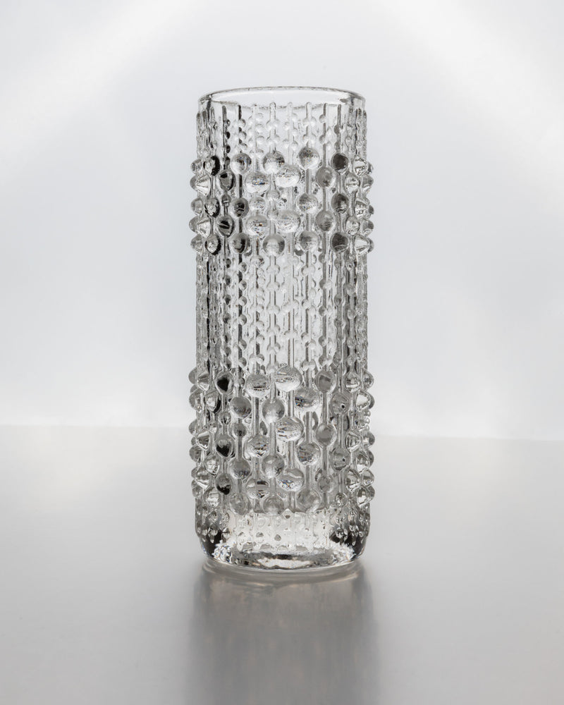 The Glass Vase by Frantisek Peceny for Bottega Jacobs (1965) is a tall, clear vintage piece with a cylindrical shape and raised droplet texture, displayed on a reflective surface against a plain light background.