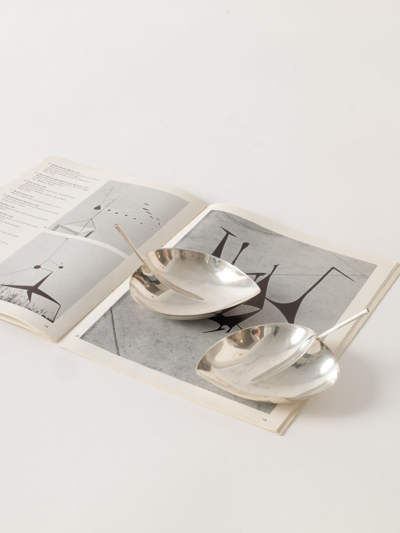 An open book with black and white images laid on a white surface. Two shiny, silver, leaf-shaped Tapio Wirkkala Silver Bowls from Collection apart are placed on top of the book. Each bowl contains a single white-handled spoon. The background is minimalistic and clean.