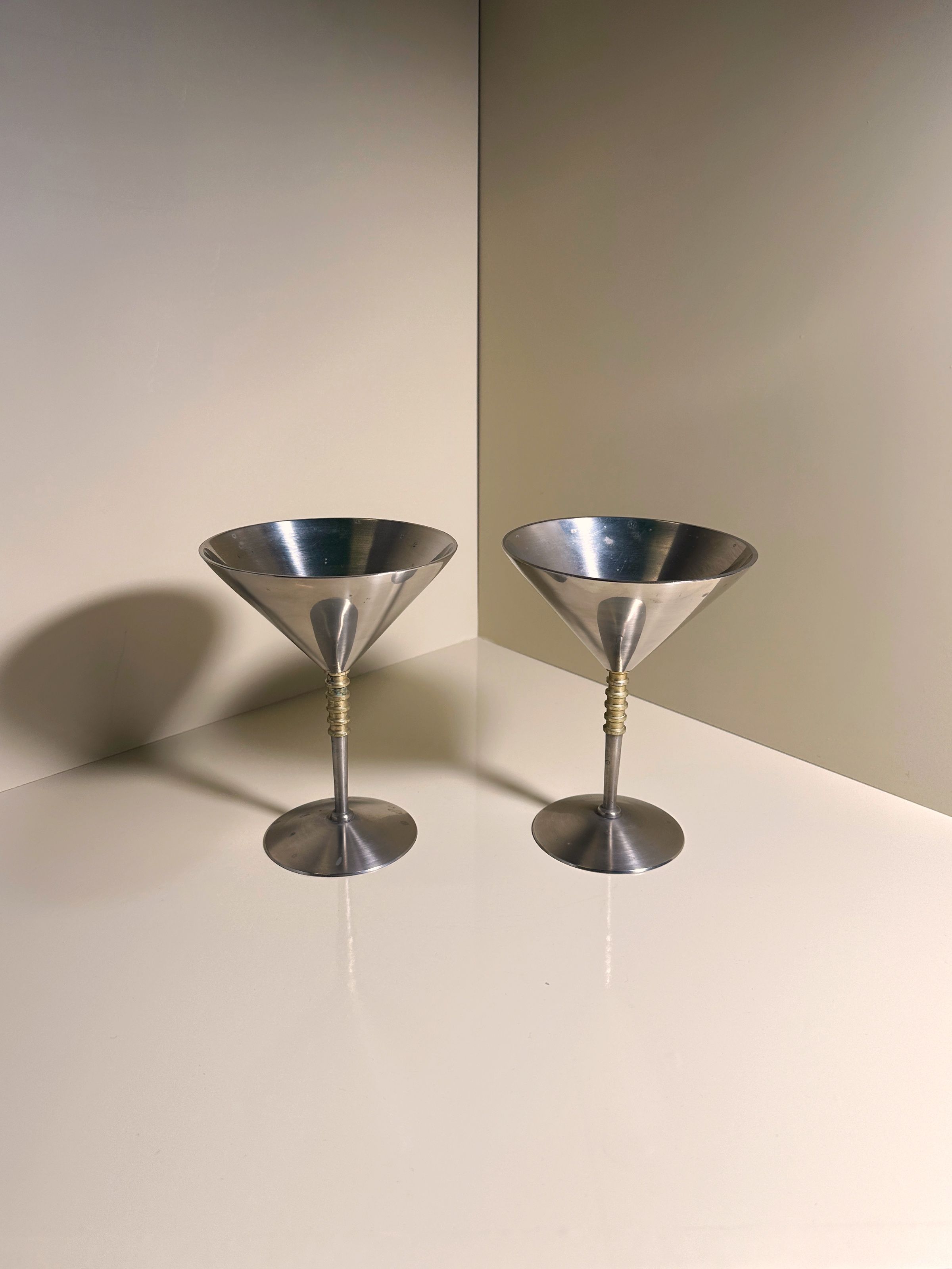 A pair of Dodo Vintage solid metal coupés feature brass detail stems, set against a light beige background. Their smooth stainless steel surfaces reflect light to create subtle highlights.