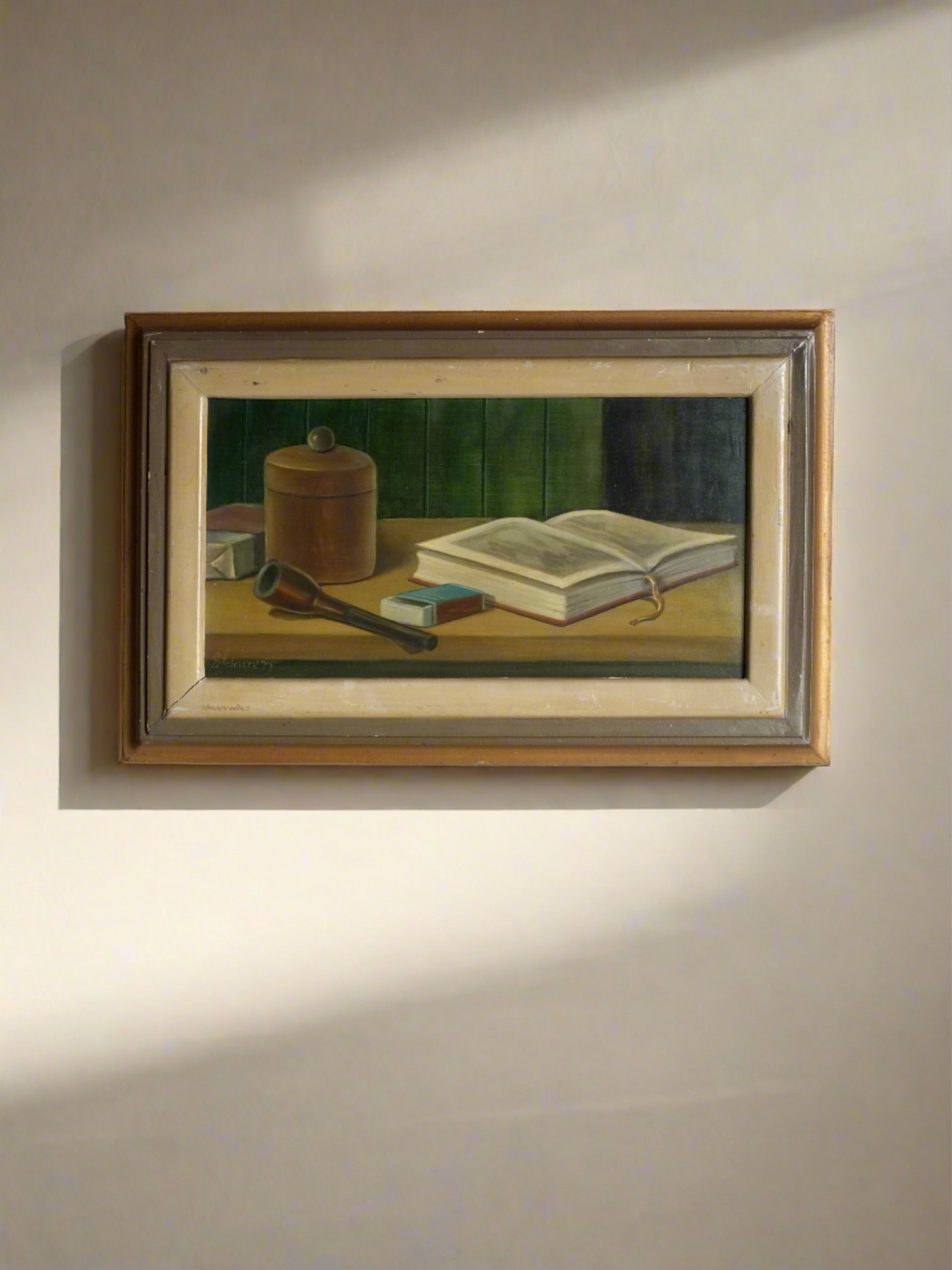 A framed Vintage Realism Still Life Oil Painting | 1970s by Veter Vintage hangs on a plain, grey wall. The artwork, of European origin, depicts a brown cylindrical container, a pipe, matchbox, and an open book placed on a wooden surface against a green background—all in excellent vintage condition.